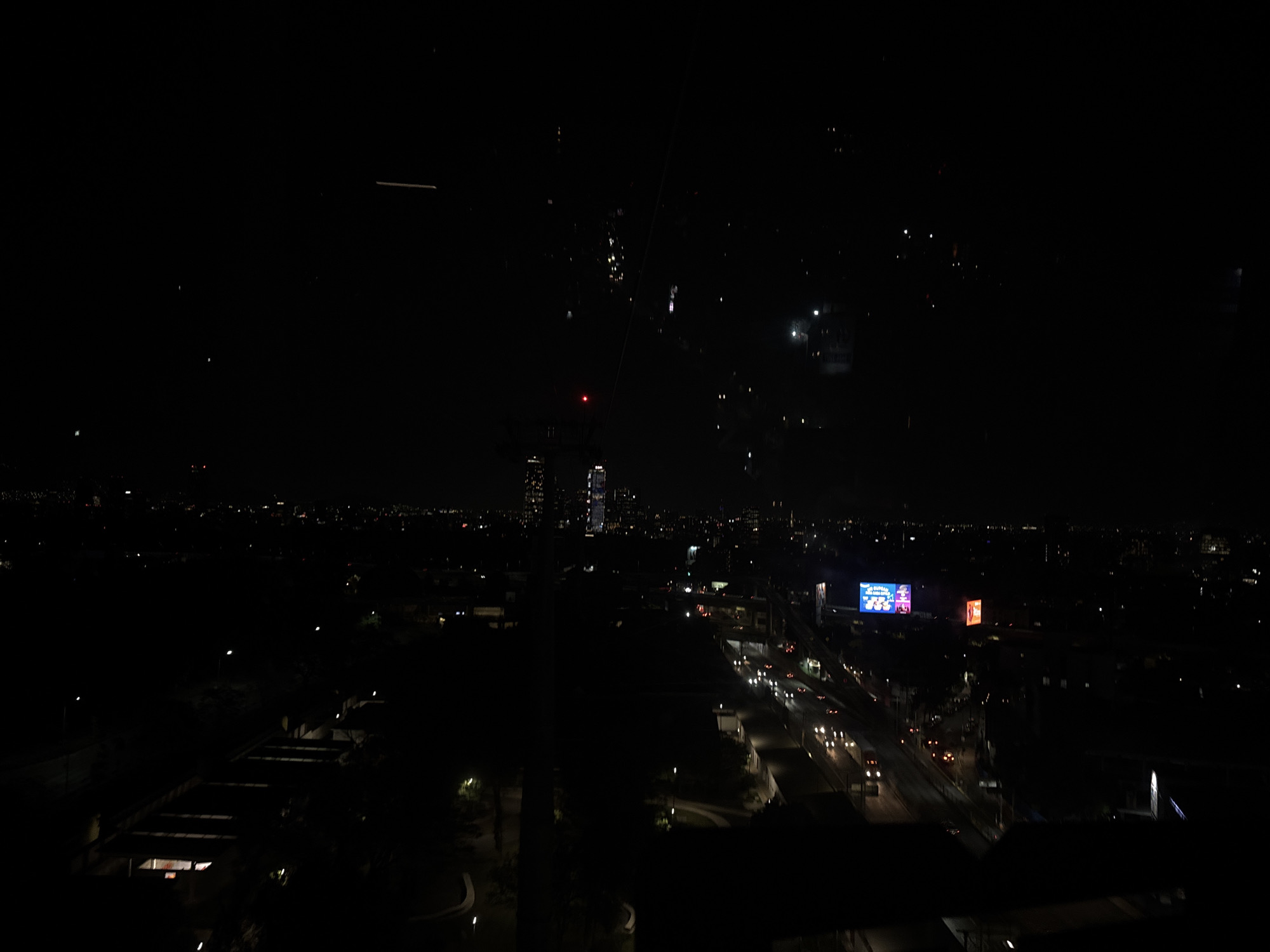 The image shows a nighttime cityscape view from a high vantage point, likely a building window. The location is Mexico City, Mexico, as evidenced by the cityscape and the prominent billboard. In the distance, a cluster of taller buildings can be seen. Below, a network of roads is visible, with moving vehicle lights streaking through the scene. Some buildings are seen at the bottom. Streetlights are intermittently illuminating the urban landscape. A large billboard displaying illuminated graphics and text can be seen with another billboard to the right of it. The overall scene is dimly lit, with the brightest points being lights in buildings and on vehicles.