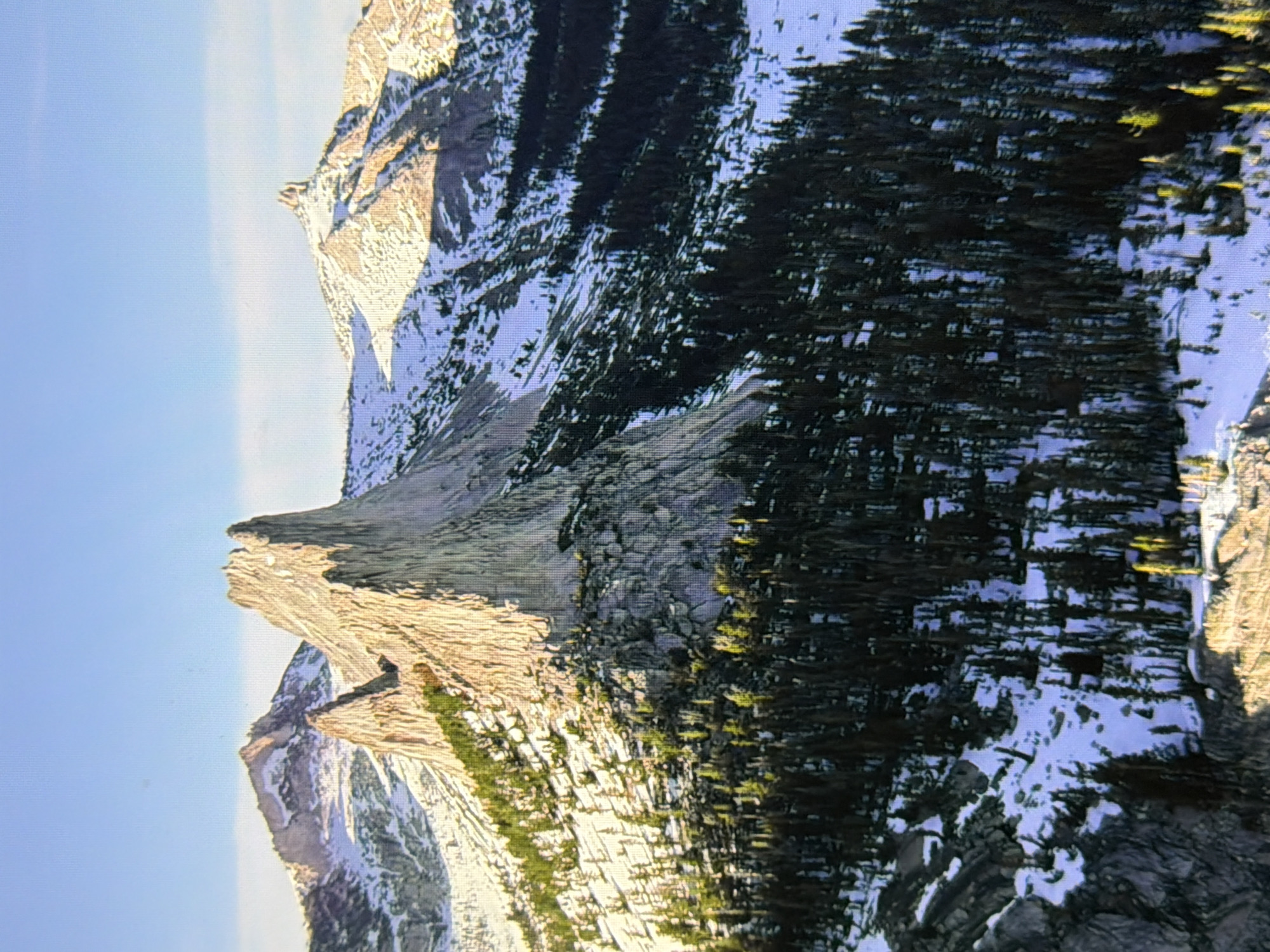 The image depicts a rugged mountain range with snow-capped peaks and coniferous trees. The sunlight casts long shadows, highlighting the texture of the rocky terrain. The sky is clear and blue. The scene is devoid of any human presence or urban elements, suggesting a remote wilderness location. There is no discernible connection to the city of Kyiv, Ukraine.