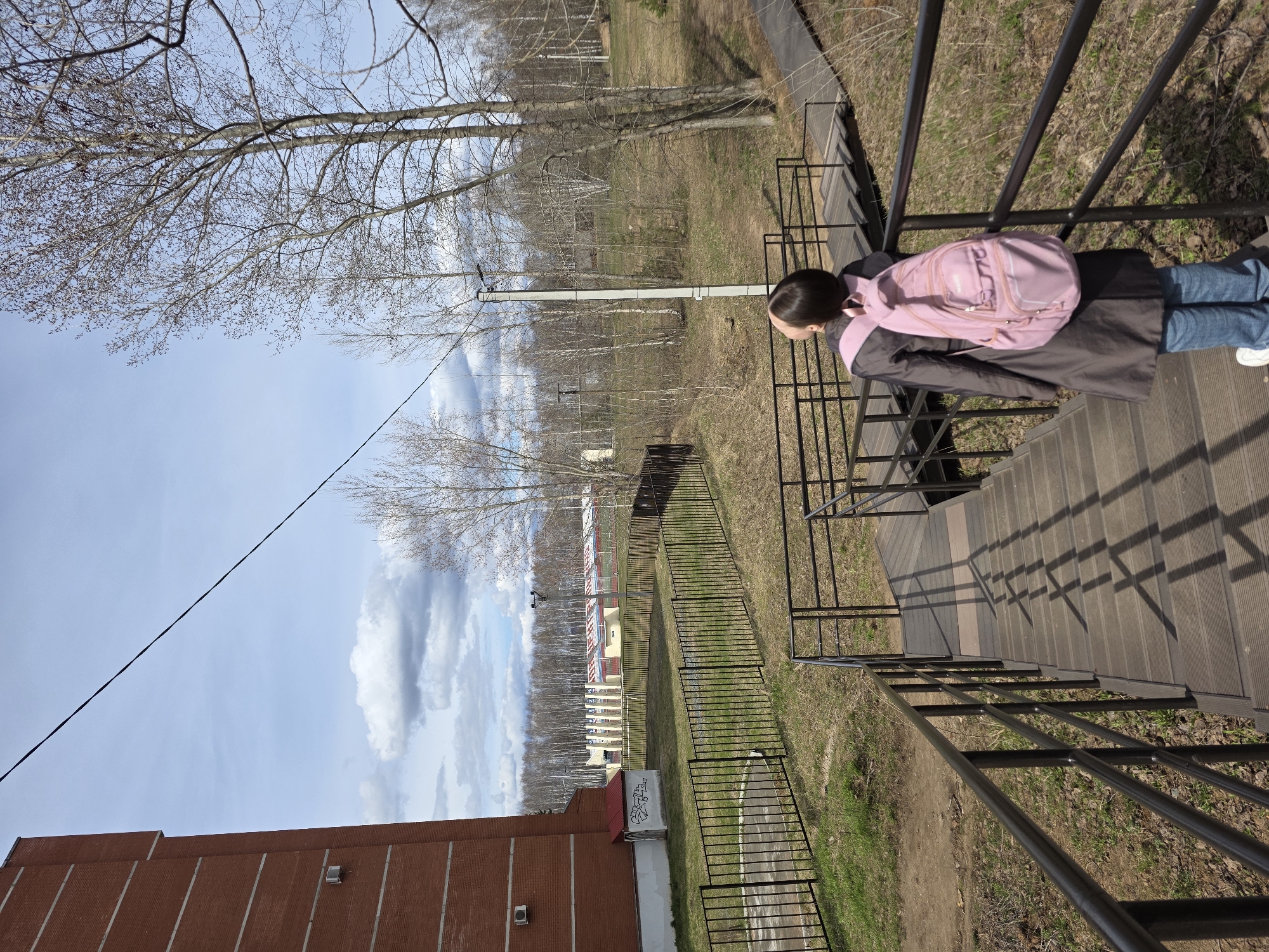 This outdoor image, captured in Chaikovsky, Russia, features a person standing on a wooden-look staircase with black metal railings, looking down into the scene below. The individual is seen from behind, wearing a dark coat, blue jeans, white sneakers, and a prominent pink backpack. Their dark hair is pulled back. Shadows cast by the railings on the steps suggest bright, sunny daytime conditions.
To the left of the stairs, a metal fence runs parallel, separating the immediate area from a grassy field dotted with bare deciduous trees, indicative of early spring or late autumn. A utility pole with wires stands in the mid-ground.
Further in the distance, a complex of buildings is visible, including what appears to be a stadium or sports facility with distinctive seating rows. A sign near this complex reads "ЗАРЯ" (Zarya) and "СПОРТИВНЫЙ КОМПЛЕКС" (Sports Complex), clearly identifying it as a sports venue.
On the far left of the frame, a large red brick building with vertical white lines and a small air conditioning unit dominates the foreground. A smaller white structure with a red roof sits beneath it, displaying some illegible graffiti.
The sky above is bright blue with scattered white cumulus clouds. A power line diagonally crosses the upper left portion of the sky. The overall scene depicts a calm day in an area that combines natural elements with urban infrastructure and a sports complex.