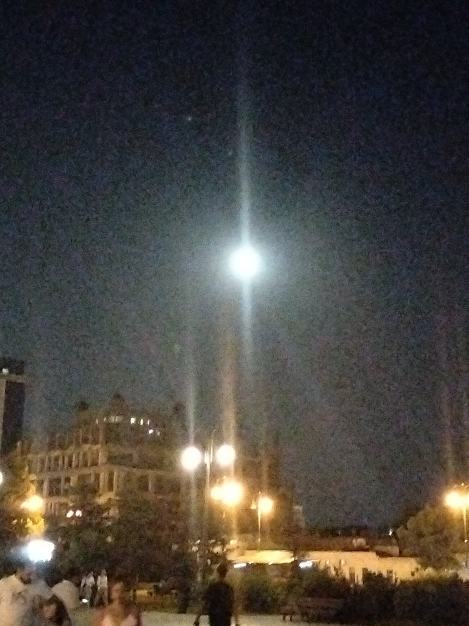 The image depicts a night scene in Baku, Azerbaijan, likely near the seaside boulevard. A bright moon or a strong light source dominates the upper portion of the frame, casting a glare that obscures details. Below this, a multi-story building with numerous illuminated windows is visible, suggesting it is either an apartment complex or a hotel. Several streetlights with warm, yellow illumination are scattered throughout the mid-ground and right side of the image. In the foreground, several figures are present, some of whom are partially obscured and appear to be walking or standing. The overall impression is of a lively urban environment at night, with artificial lights and natural moonlight contributing to the illumination.
