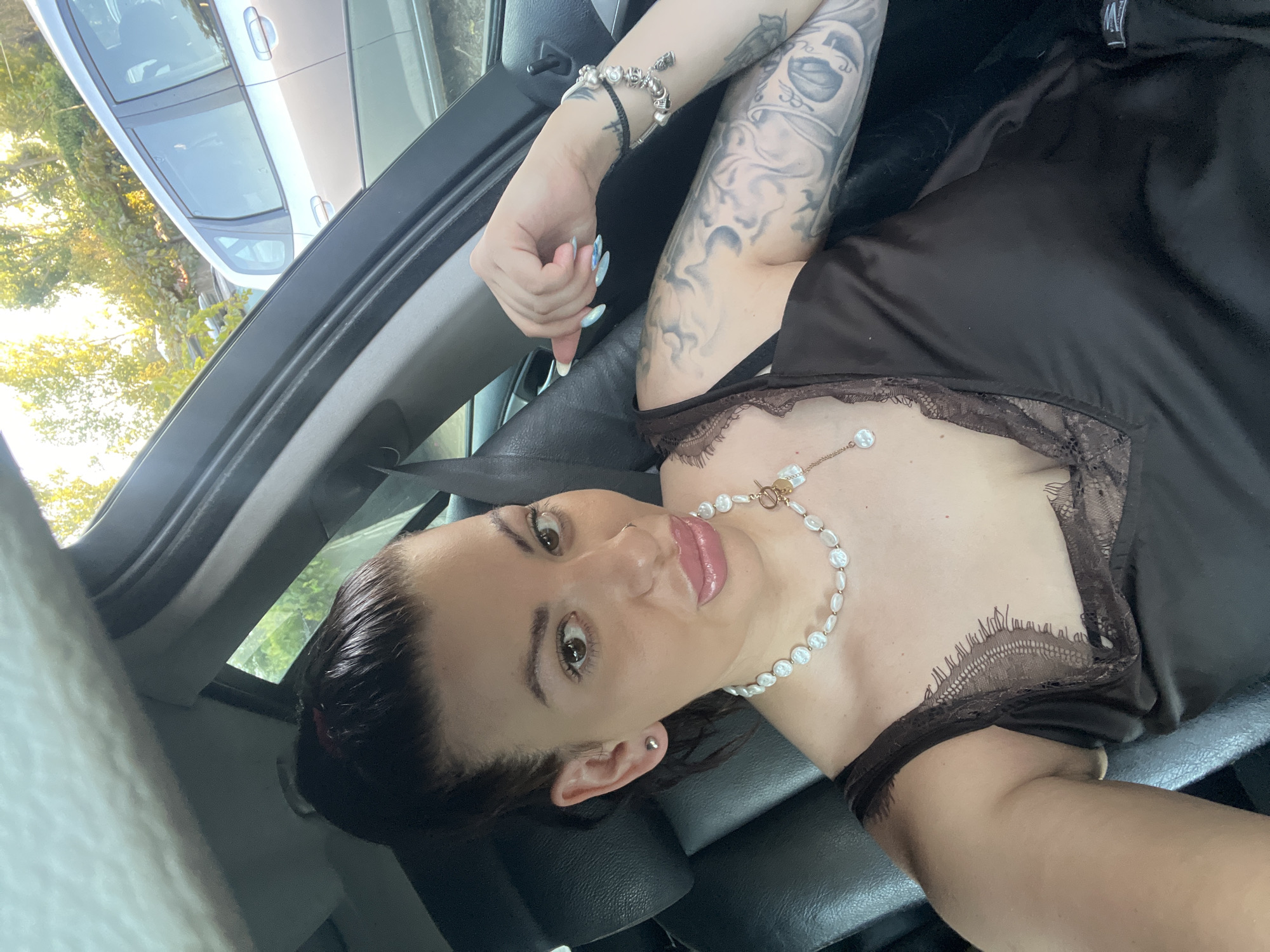 The image depicts a woman in the driver's seat of a car, parked outdoors. She is looking at the camera and posing with her hand raised. The woman has dark hair pulled back in a ponytail, is wearing a brown satin top with lace trim, and a pearl necklace. She has tattoos on her arm and wrist. The background shows trees and other cars, suggesting an urban or suburban setting. The image was taken in Sozopol, Bulgaria.