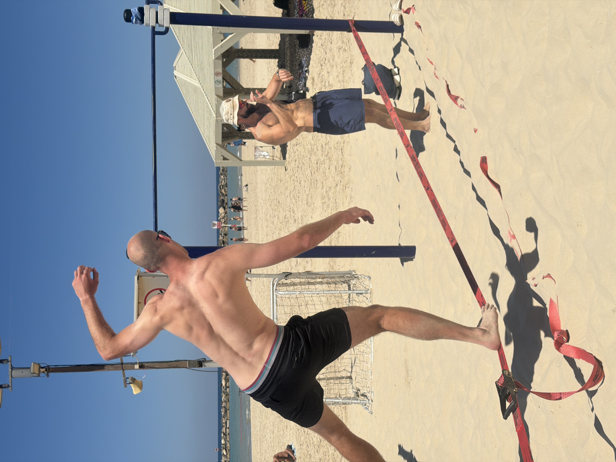 This photograph captures a bright, sunny scene on a beach in Tel Aviv, Israel, likely during midday or early afternoon given the strong, distinct shadows. The image is composed of two men engaging in slacklining on the sand, with the clear blue sky above and the Mediterranean Sea in the distance.
In the foreground, a shirtless man with a bald head and black shorts is attempting to balance on a red slackline. His left leg is extended forward, and his right foot is placed on the tensioned strap. His back muscles are well-defined as he maintains his posture. Sunglasses rest on the back of his head.
Further back, to the right, another shirtless man with a beard, wearing a white hat and sunglasses, stands watching. He wears dark blue shorts and has his hands slightly raised, appearing to either be observing the slackliner intently or perhaps giving instructions.
The slackline itself is red, with a black ratchet mechanism visible on the right side, anchoring it to the sand. Visible text on the slackline includes "BEACHLINER" and "SLACKLINE TOOLS".
The beach is sandy, and in the background, there is a light-colored wooden canopy structure with slatted sides, providing shade. A tall blue pole, possibly part of the beach's infrastructure, extends upwards. In the distance, people can be seen walking along the shore near the water, which is separated from the beach by a line of rocks. A metal gate or barrier is also visible on the sand. The overall impression is one of leisure and outdoor activity on a beautiful day.