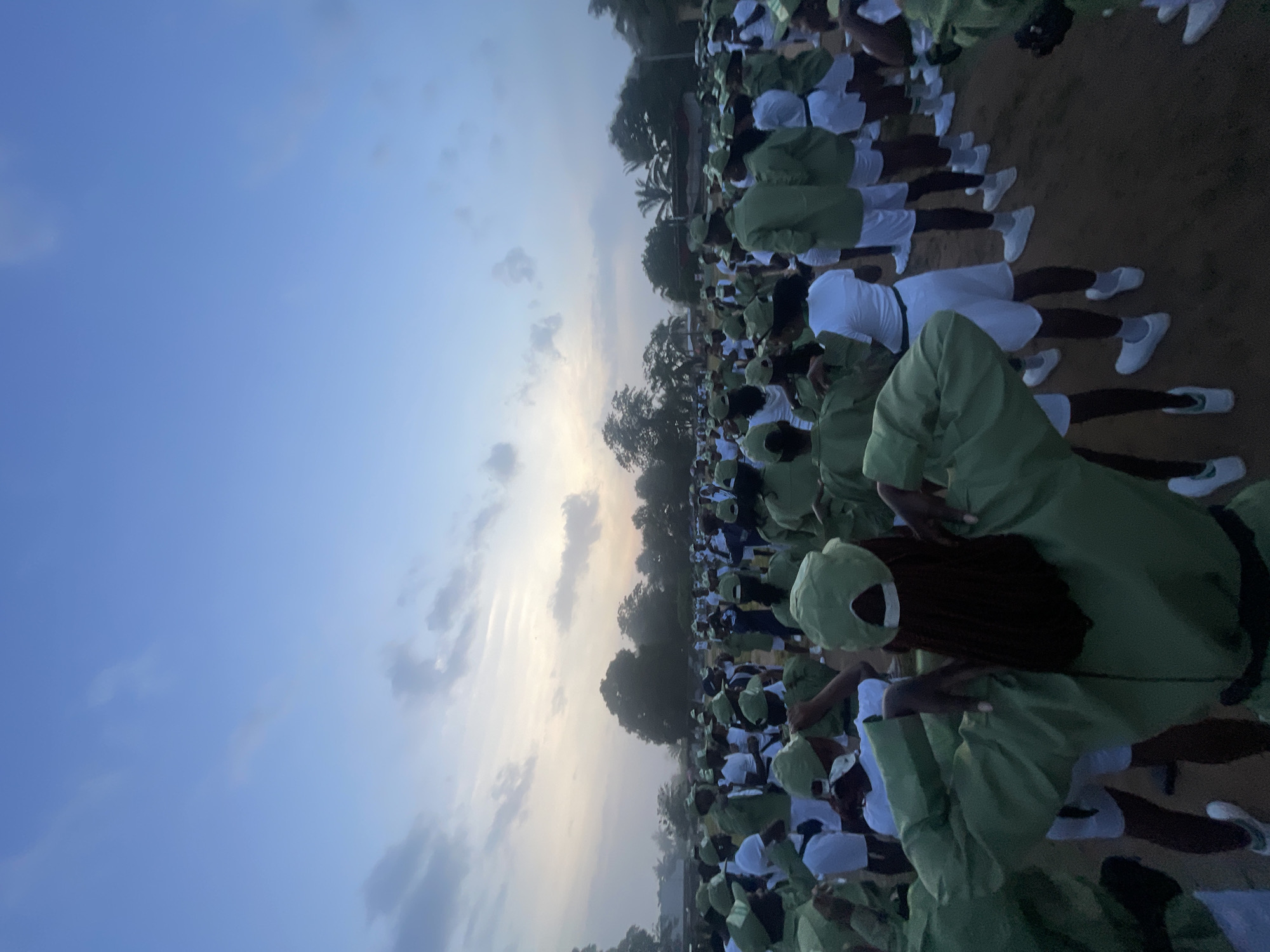 In the city of Ise-Ekiti, Nigeria, a large group of young adults, predominantly women, are gathered outdoors for what appears to be a morning assembly or orientation. They are all dressed in matching khaki uniforms consisting of green shirts, white shorts, and green caps, with white socks and white sneakers. The scene is captured at dawn or dusk, with the sky displaying soft hues of blue and scattered clouds, illuminated by the rising or setting sun.
The individuals are arranged in a field, with a scattering of trees visible in the background, suggesting a rural or semi-urban setting. Some are standing in rows, while others are in more informal clusters, facing in various directions. The general atmosphere appears to be one of order and collective activity, possibly related to a national youth service or a similar program. The low lighting indicates it's either early morning or late evening, contributing to the diffused, soft quality of the light. The ground is a dirt or gravel surface.