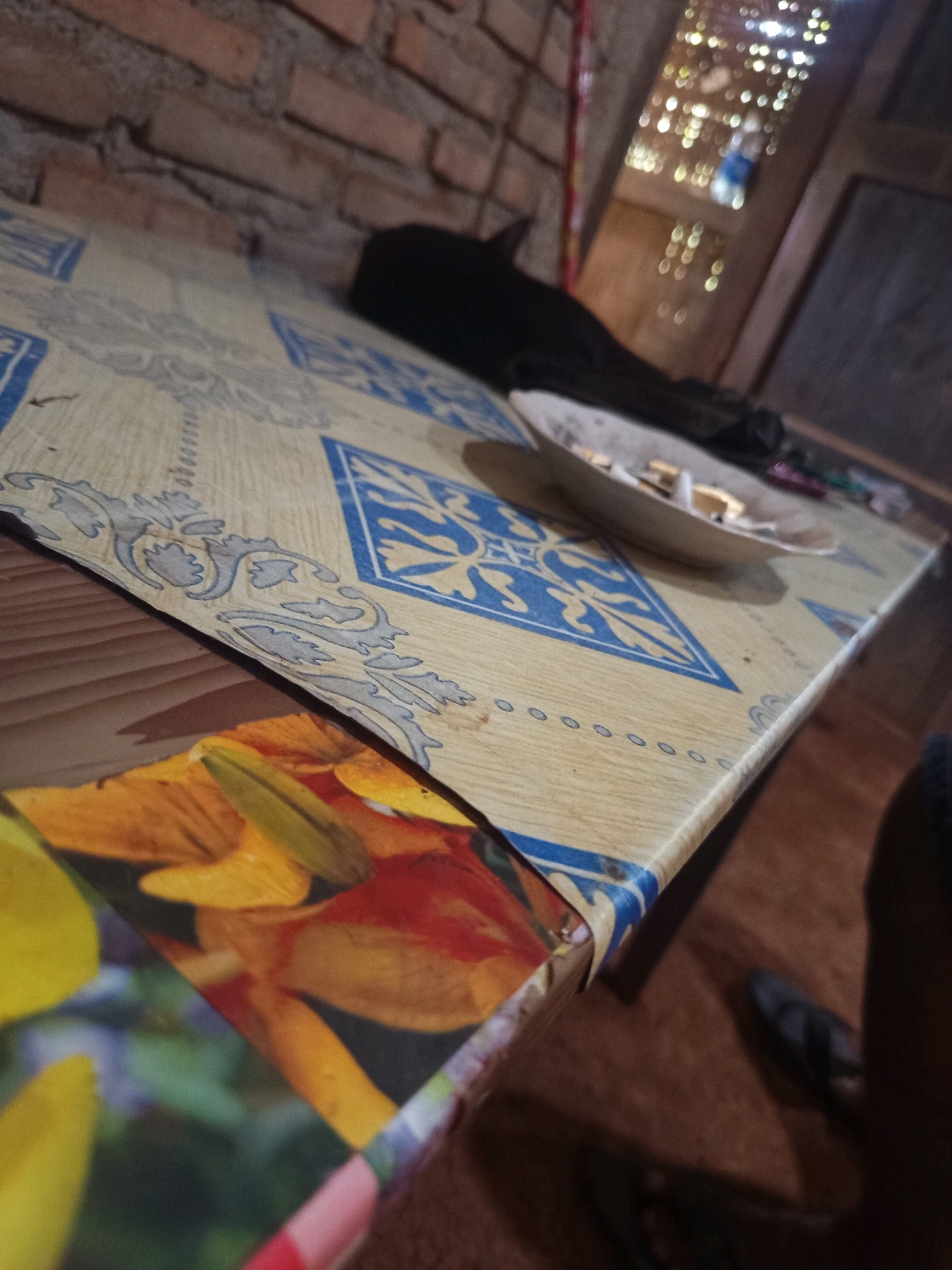 The image depicts a table with a patterned cloth covering it. The cloth has a beige base with blue geometric designs and floral motifs. On the table, there is a small white plate with cigarette butts and possibly some wrappers. A black object, which could be a piece of cloth or an animal, is partially visible on the table. In the background, a brick wall is visible, and to the right, a blurred out window shows lights reflecting, suggesting an indoor setting at night. The floor appears to be made of wood. A pair of dark-colored flip-flops are on the floor near the edge of the table. The overall scene suggests a casual, possibly domestic or outdoor seating area. The context of Karang Sari, Indonesia, implies a tropical or subtropical climate, though the image itself doesn't provide explicit visual cues to confirm this.