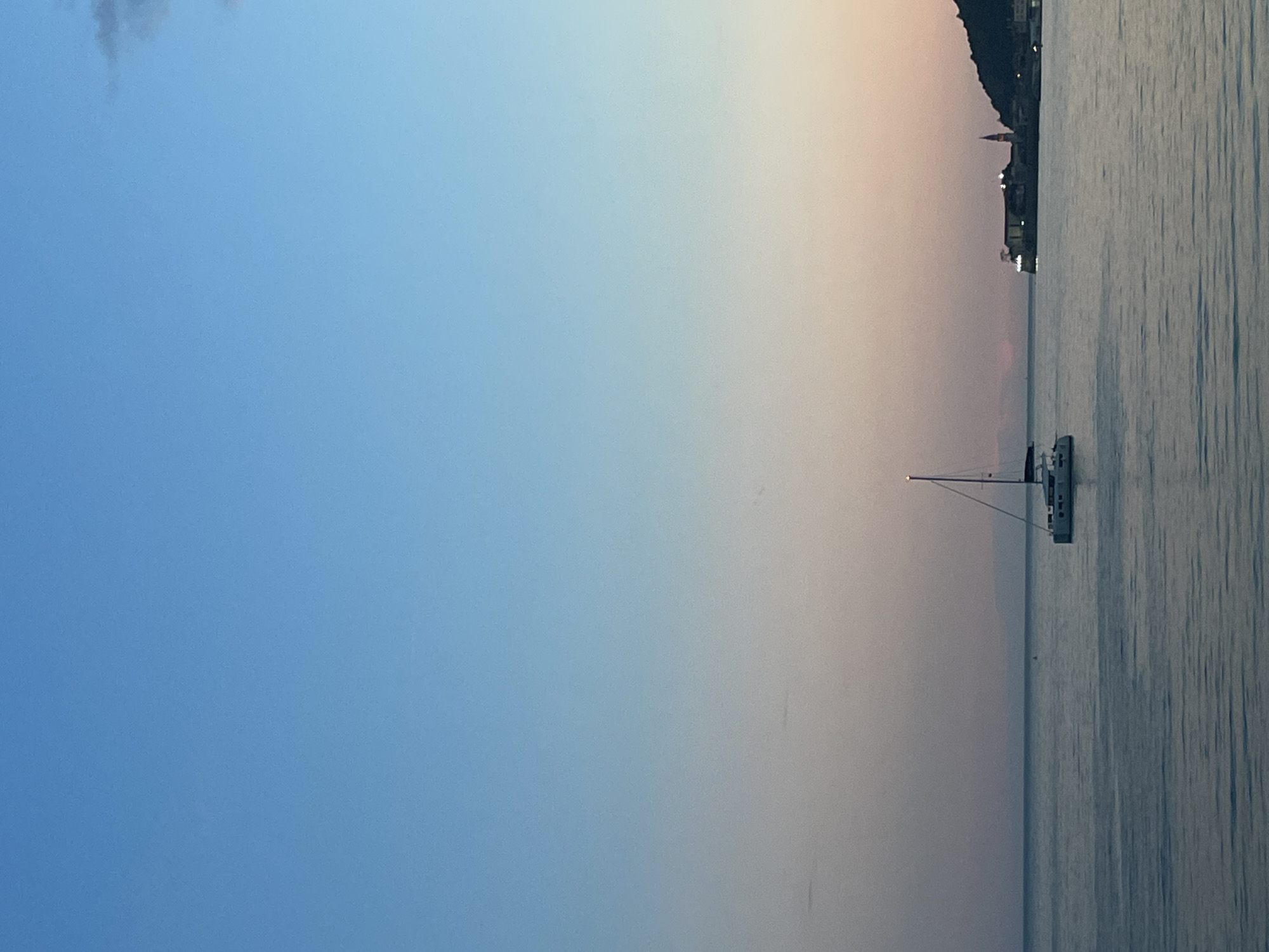 The image shows a body of water at dusk, possibly the Adriatic Sea, with a sailboat anchored near the shoreline. On the right side of the frame, a coastline with buildings, including what may be a church steeple, is visible. The buildings and the angle of the land suggest this could be in or near the city of Budva, Montenegro. The sky transitions from a light blue on the left to a peachy, orange color near the water. The water is a dark color, reflecting the fading light of the sky. There is no visible human activity except for the presence of the boat.