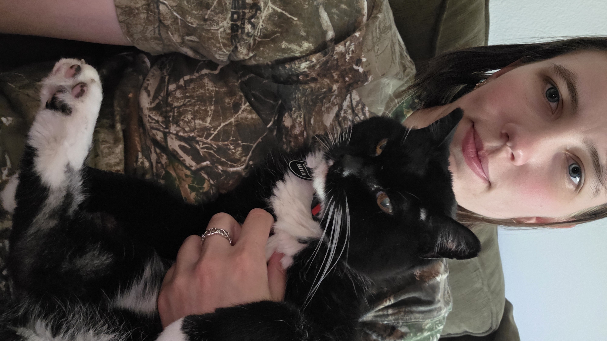 A woman with blue eyes and brown hair is holding a black and white cat. She is wearing a camouflage shirt. The cat has a white chest and white paws. The woman is wearing a ring on her finger. The image appears to have been taken indoors, possibly in a home. The background shows a couch and a wall. The location is not specified in the provided text or image.
