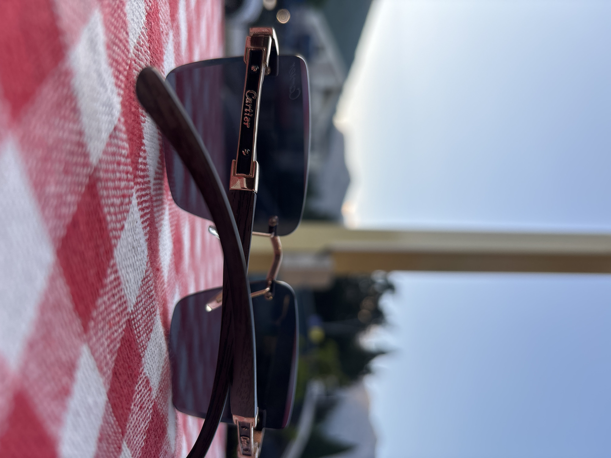 The image shows a close-up view of a pair of Cartier sunglasses resting on a red and white checkered tablecloth. The sunglasses have dark lenses and a rose gold frame with black accents. The "Cartier" logo is visible on the temple. In the background, out of focus, are trees, buildings, and mountains under a clear blue sky, suggesting an outdoor setting, possibly in a mountainous coastal region like Budva, Montenegro. The lighting indicates it is daytime, likely late afternoon or early evening due to the soft light. There are no people visible in the frame.