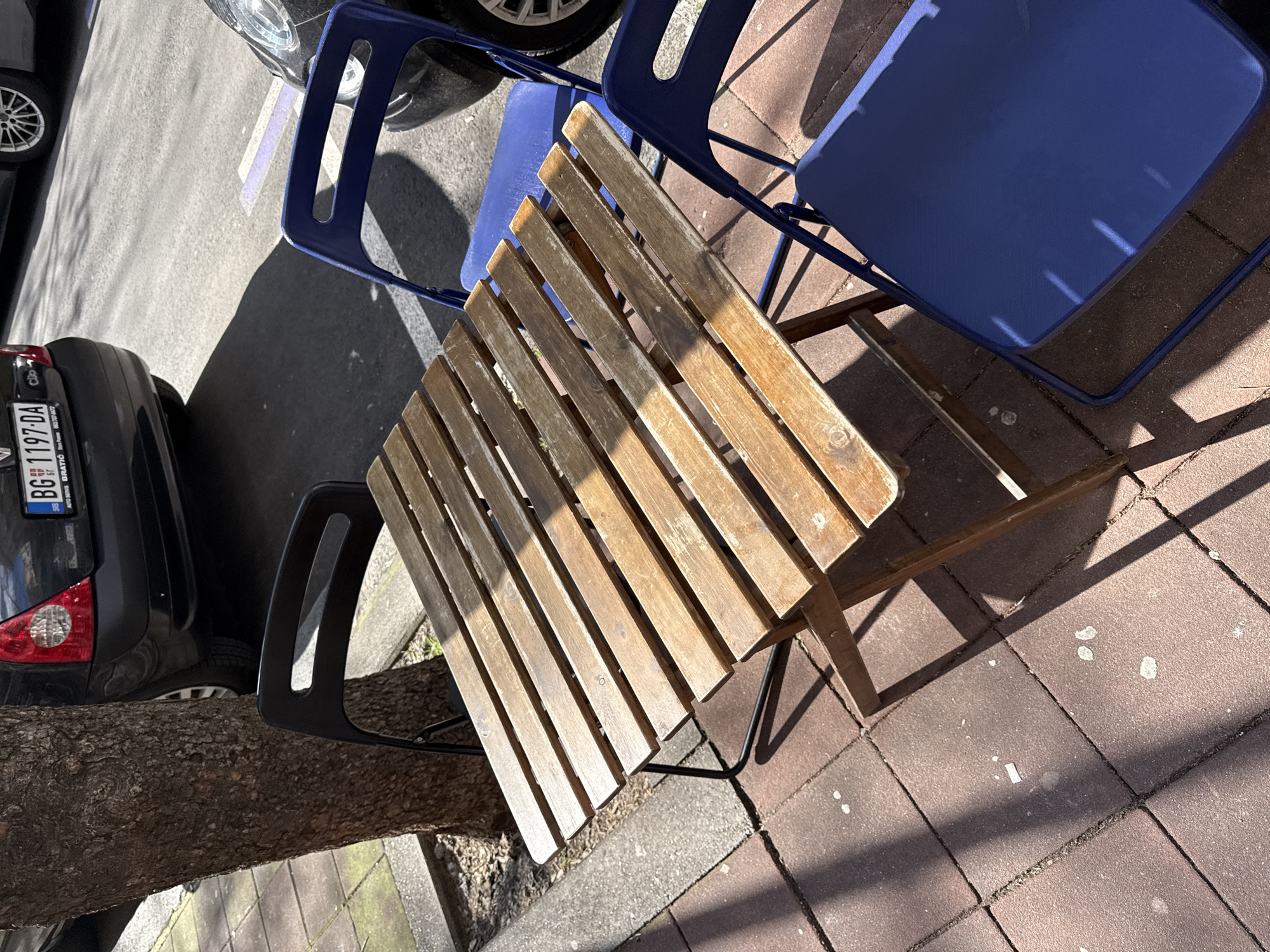 The image presents a top-down view of a small, outdoor seating area. A square wooden table with a slatted surface is centrally located. Two blue metal chairs flank the table, one positioned to the upper right and another to the upper left. A black metal chair is located to the left of the table. To the left of the black metal chair is a tree trunk. Partially visible on the left side of the image is a dark-colored car, specifically its rear portion, with the license plate 'BG 1197-DA' displayed, indicating Belgrade, Serbia. The ground beneath the seating arrangement transitions from asphalt near the car to red brick paving. The area appears to be outdoors on a sunny day.