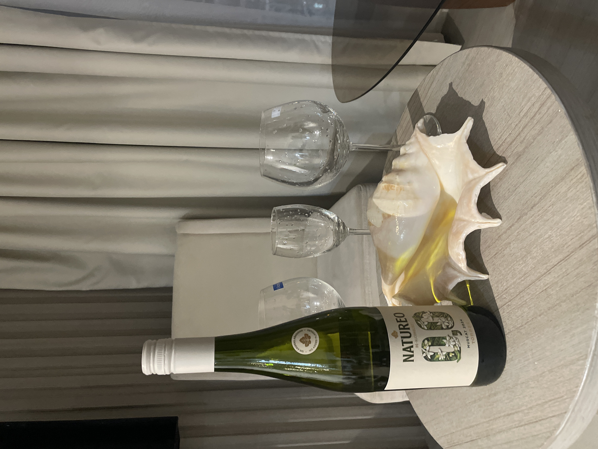 On a round, light-wood-grain-surface table in a room, there is a bottle of Natureo de-alcoholized Muscat wine, two empty wine glasses, and a large conch shell. The wine bottle has a white label and a white screw-top cap. The background features a light-colored, draped curtain. The scene suggests an indoor setting, possibly in Bắc Nha Trang, Vietnam. There are no people present in the image.