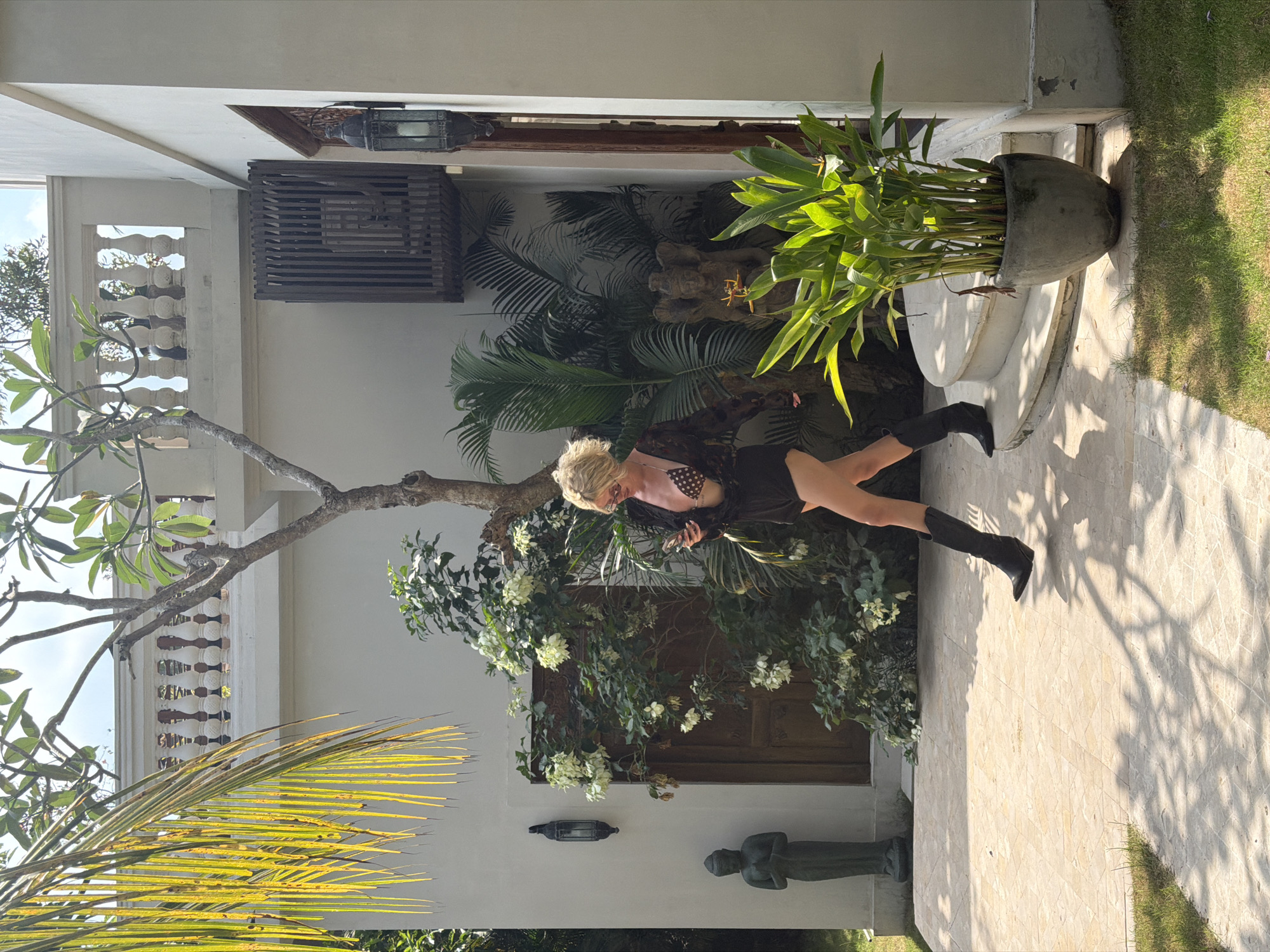 A blonde woman in a polka-dot bikini top and black shorts, wearing a sheer black kimono and black cowboy boots, walks across a tan tiled patio. She holds a smartphone in her right hand. She is positioned in the mid-right of the frame. Behind her, to the left, is a weathered wooden door, and to the right of the door is lush green foliage with white flowers. A dark green statue of a meditating figure stands near the door. A frangipani tree with a thin trunk is visible in the upper half of the frame, with a railing and balustrade of a building visible behind it. To the right of the woman, a large potted plant with green leaves sits next to a curved concrete staircase. The setting appears to be an outdoor courtyard in a tropical environment, likely a resort or private residence in North Kuta, Indonesia, as indicated by the architecture and lush vegetation. The lighting suggests it is daytime, with strong sunlight casting shadows on the patio.