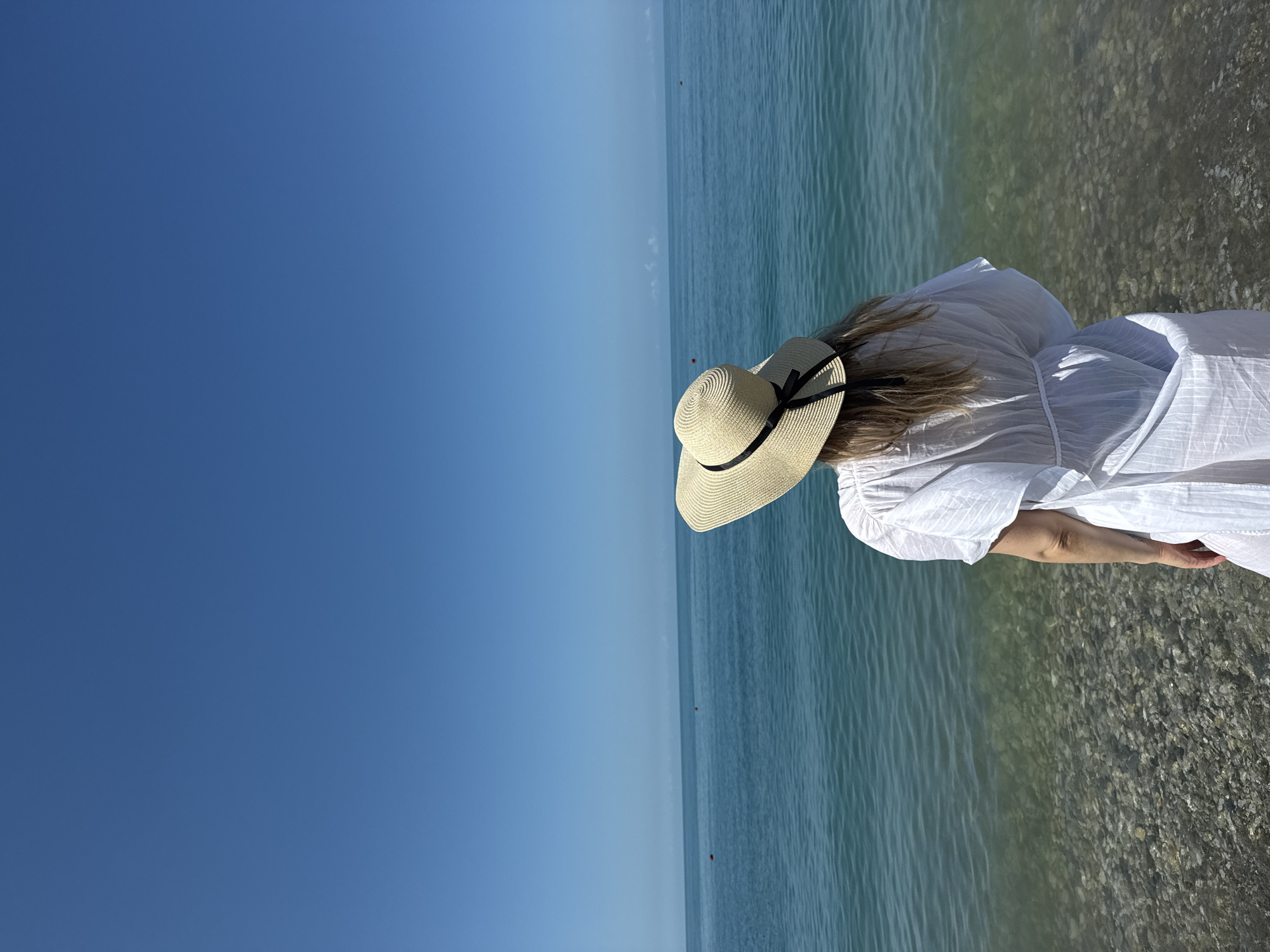 A person wearing a wide-brimmed straw hat with a black ribbon and a white flowing garment stands on a pebble beach, facing the calm, turquoise waters of the Black Sea under a clear blue sky. The water's edge is visible, with the pebbles submerged beneath the shallow water. In the distance, two red buoys mark the sea. The location is the coast of Nebug, Russia.