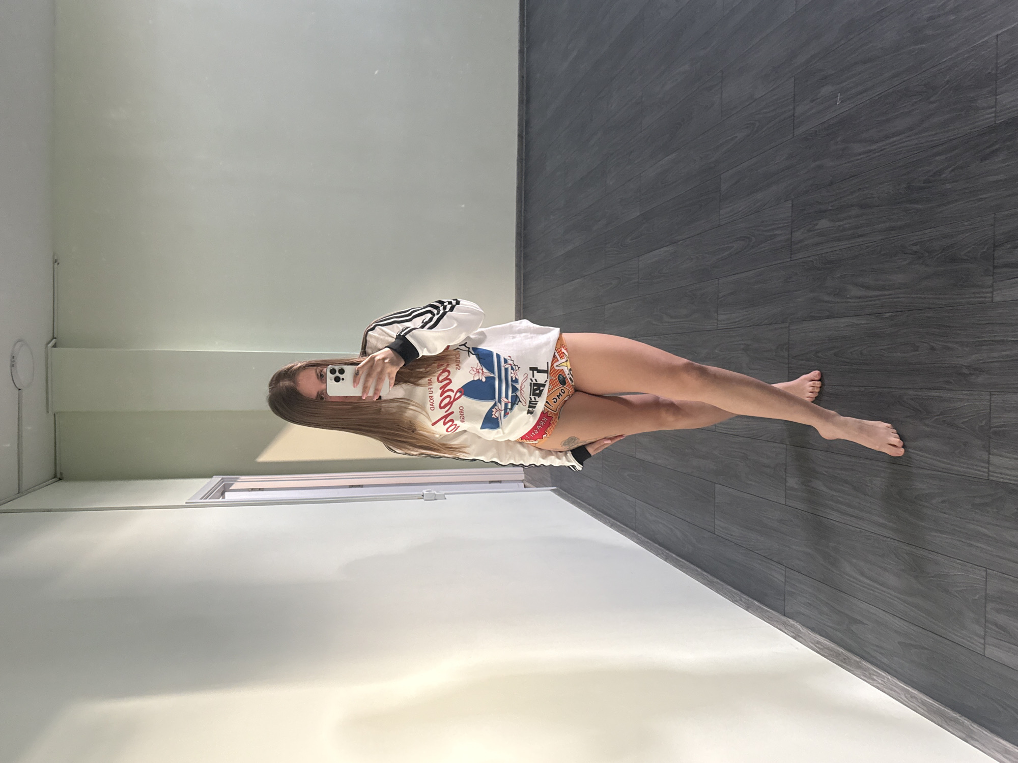 A young woman is posing for a selfie in a room, seemingly in a mirror. She is wearing a white Adidas jacket with black stripes, and a white t-shirt and patterned shorts. The room has a white wall to the left and a dark grey wood-paneled wall to the right. Based on the architecture, the room may be located in a residential building in Batumi, Georgia, but there is no way to confirm the exact location based on the available information.