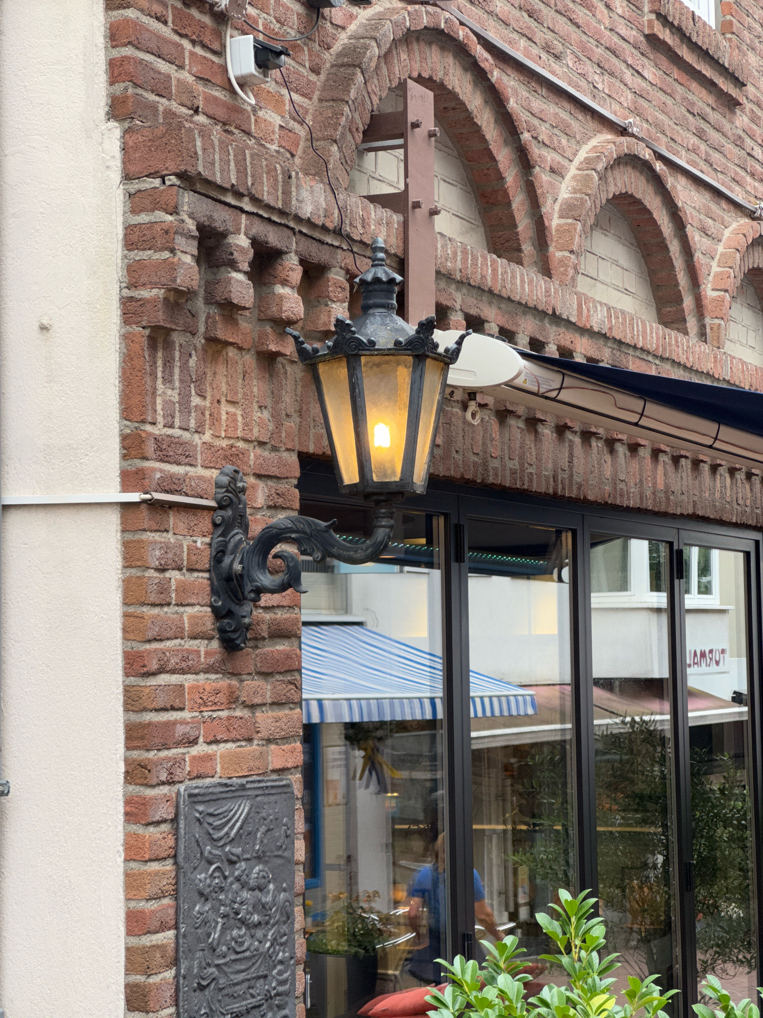 The image captures a close-up view of a building facade in the city of Cleves, Germany. The facade is constructed of red brick and features arched window openings on the upper level, lined with lighter brickwork. A black wrought-iron lamppost with an illuminated amber glass lantern is mounted on the brick wall to the left of a large glass-paneled storefront. Below the lamppost is a dark, rectangular decorative plaque affixed to the brick.
The storefront's glass reflects the street, showing a blue and white striped awning, greenery, and glimpses of interiors. A person wearing a blue shirt is visible inside the establishment, near a door. The overall scene suggests a building that combines traditional architectural elements with modern commercial space.