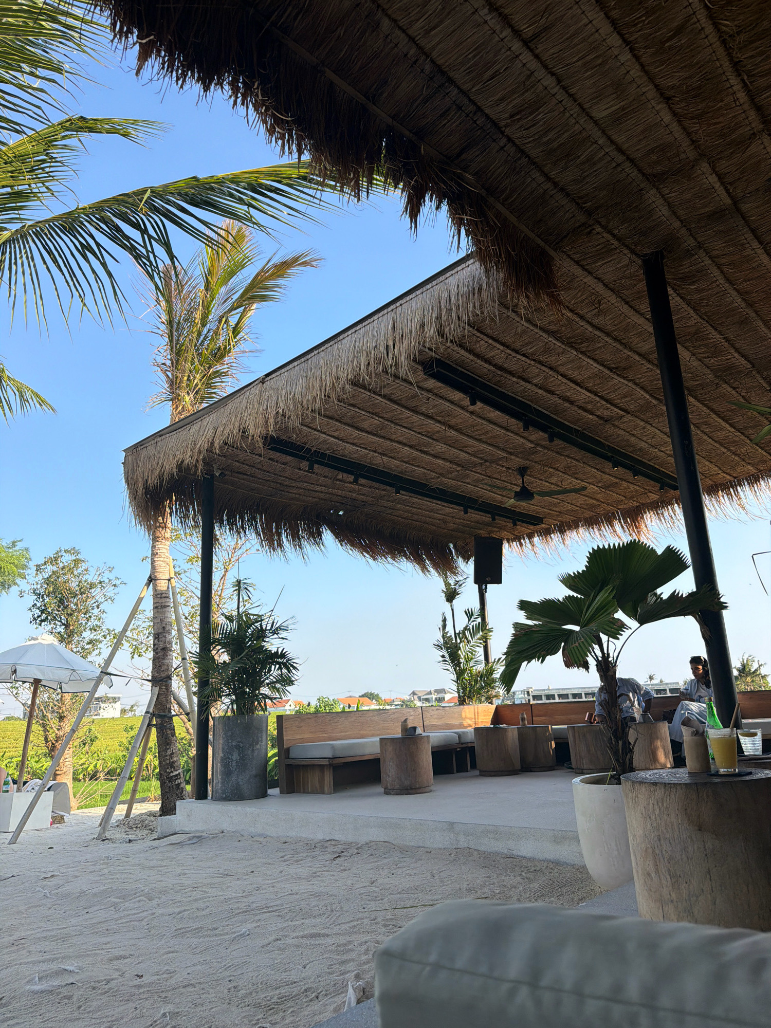 In Mengwi, Indonesia, this image captures an outdoor seating area beneath a thatched roof, illuminated by natural sunlight against a clear blue sky. Palm fronds arch over the scene, framing a view of lush greenery and distant buildings. The area features sand-covered ground and a concrete patio, furnished with wooden benches and large tree stump tables. A ceiling fan is visible beneath the thatched roof, and two individuals are seated at a table, one holding a glass of yellow liquid. A white umbrella stands to the left, and a potted palm tree adds a tropical element to the setting.