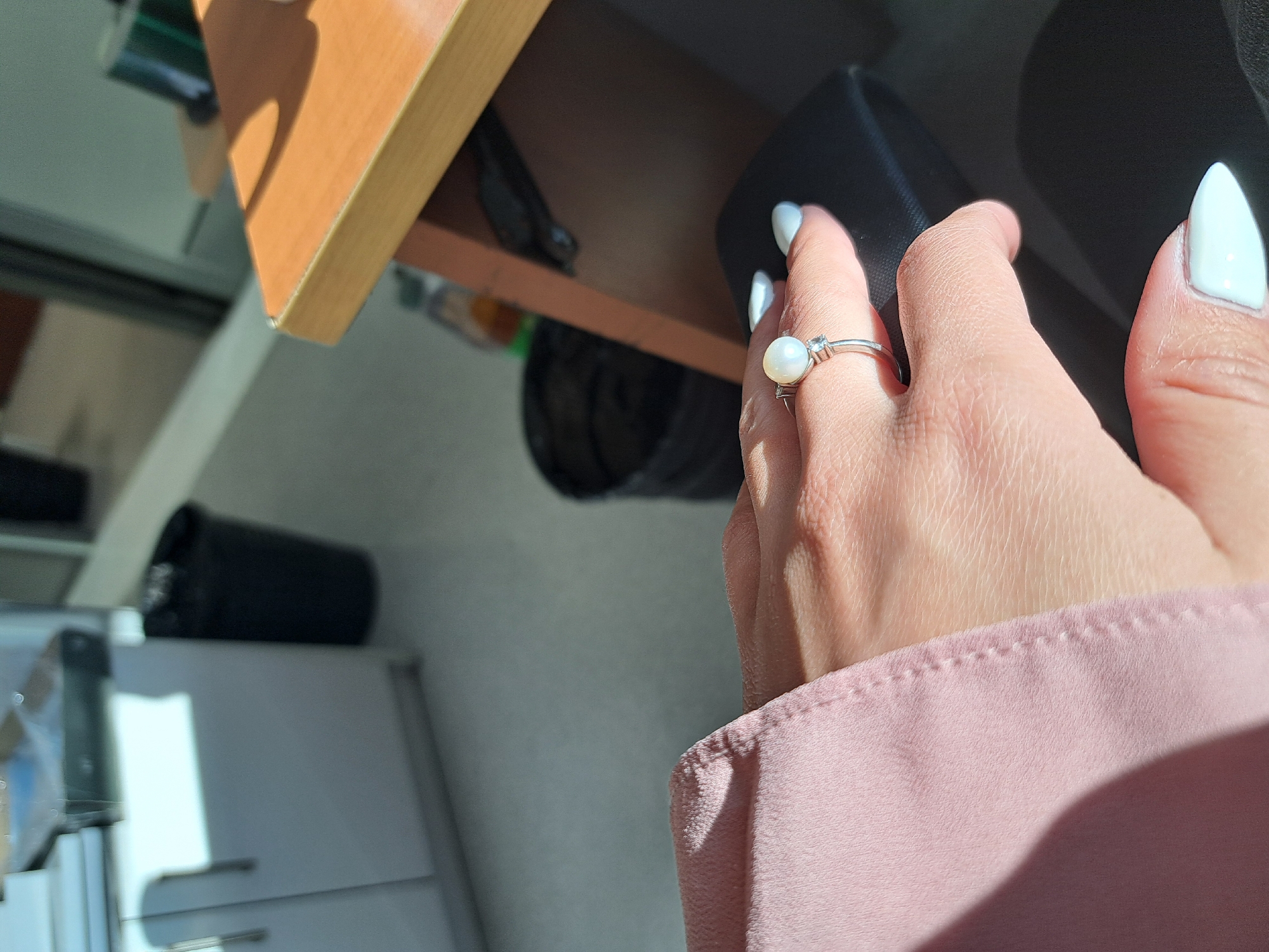 The image presents an angled, close-up view of a person's right hand in an indoor setting. The hand is adorned with a silver-toned ring featuring a prominent white pearl in the center, flanked by two small clear stones, worn on the ring finger. The fingernails are painted a glossy white and shaped into a pointed or almond style. The arm is partially visible, covered by a soft, pink garment with visible stitching along its cuff.
The hand appears to be near or holding a dark object, possibly a bag or a case, positioned under a light-brown wooden shelf or desk. Strong, direct sunlight casts sharp shadows across the wooden surface and parts of the hand, indicating a bright daytime environment. In the blurred background, a light-colored cabinet or appliance is visible to the left, and a black cylindrical object, possibly a container or plant pot, rests on a surface further back. There is no discernible specific activity taking place, nor is any text visible in the frame.