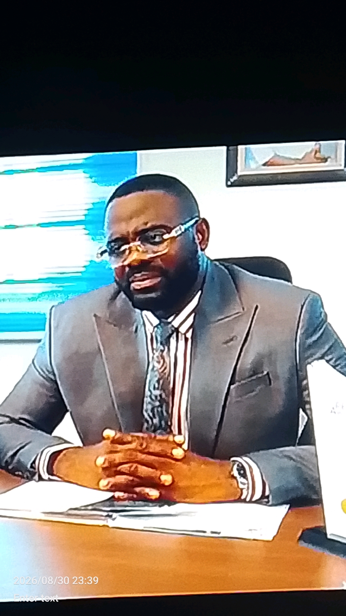 A medium shot captures a Black man wearing glasses, a suit jacket, and a tie, seated at a wooden desk. His hands are clasped together on top of a white document. He is looking forward with a neutral expression. The background features abstract artwork with blue and white tones and a framed picture. The timestamp "2026/08/30 23:39" is visible in the lower left corner. There is no discernible information in the image that would place the scene in Lokoja, Nigeria.