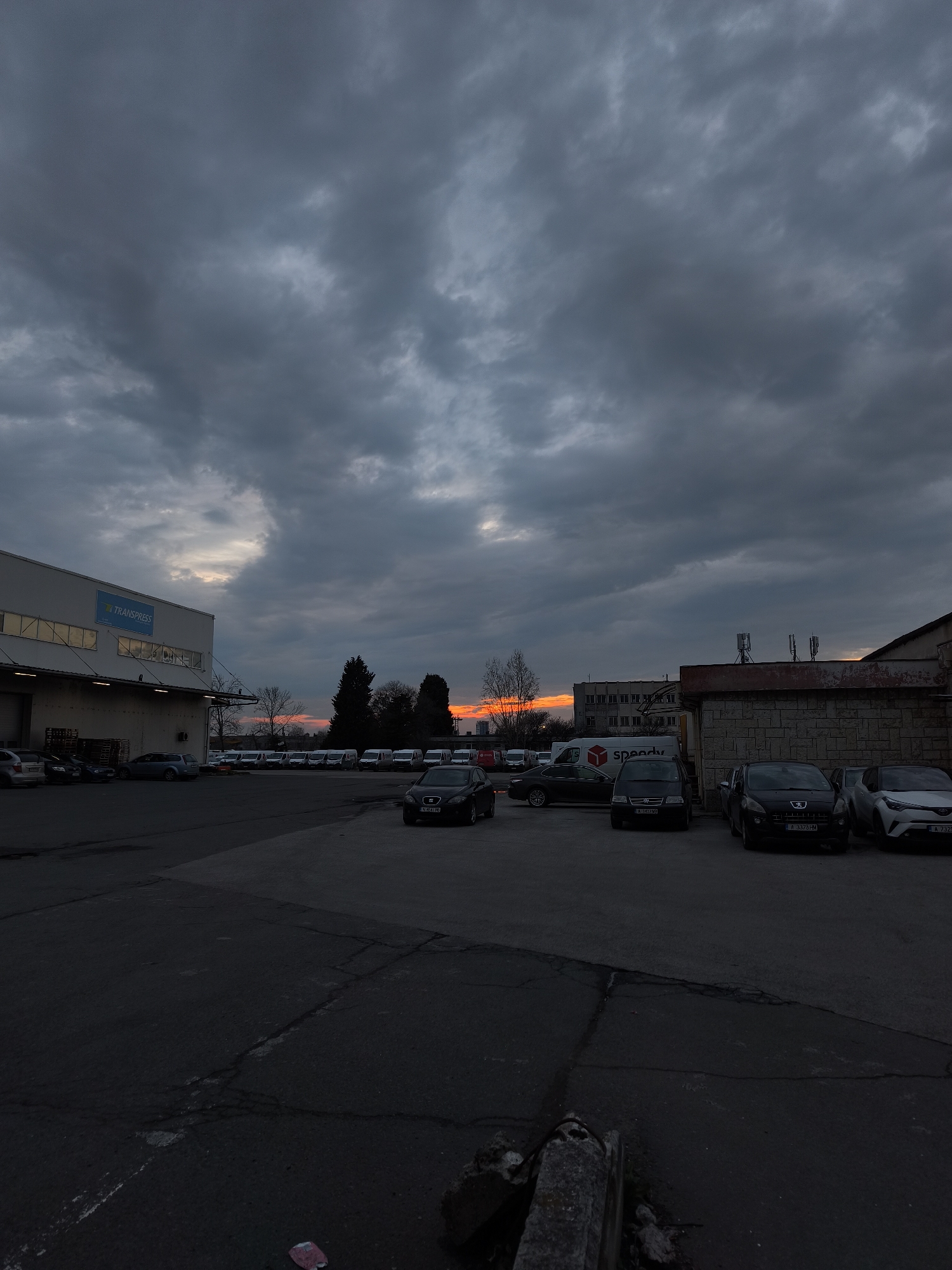 The image shows a parking lot in Burgas, Bulgaria, at dusk. The sky is heavily overcast with dark, textured clouds. A strip of orange light is visible on the horizon. A large building with "TRANSPRESS" on its facade is on the left, with several cars parked in front. A row of white vans is parked further back. To the right, more cars are parked near a building with antennas on its roof. A "Speedy" delivery van is also parked in this area. The pavement of the parking lot is cracked and uneven. The overall scene is dimly lit.