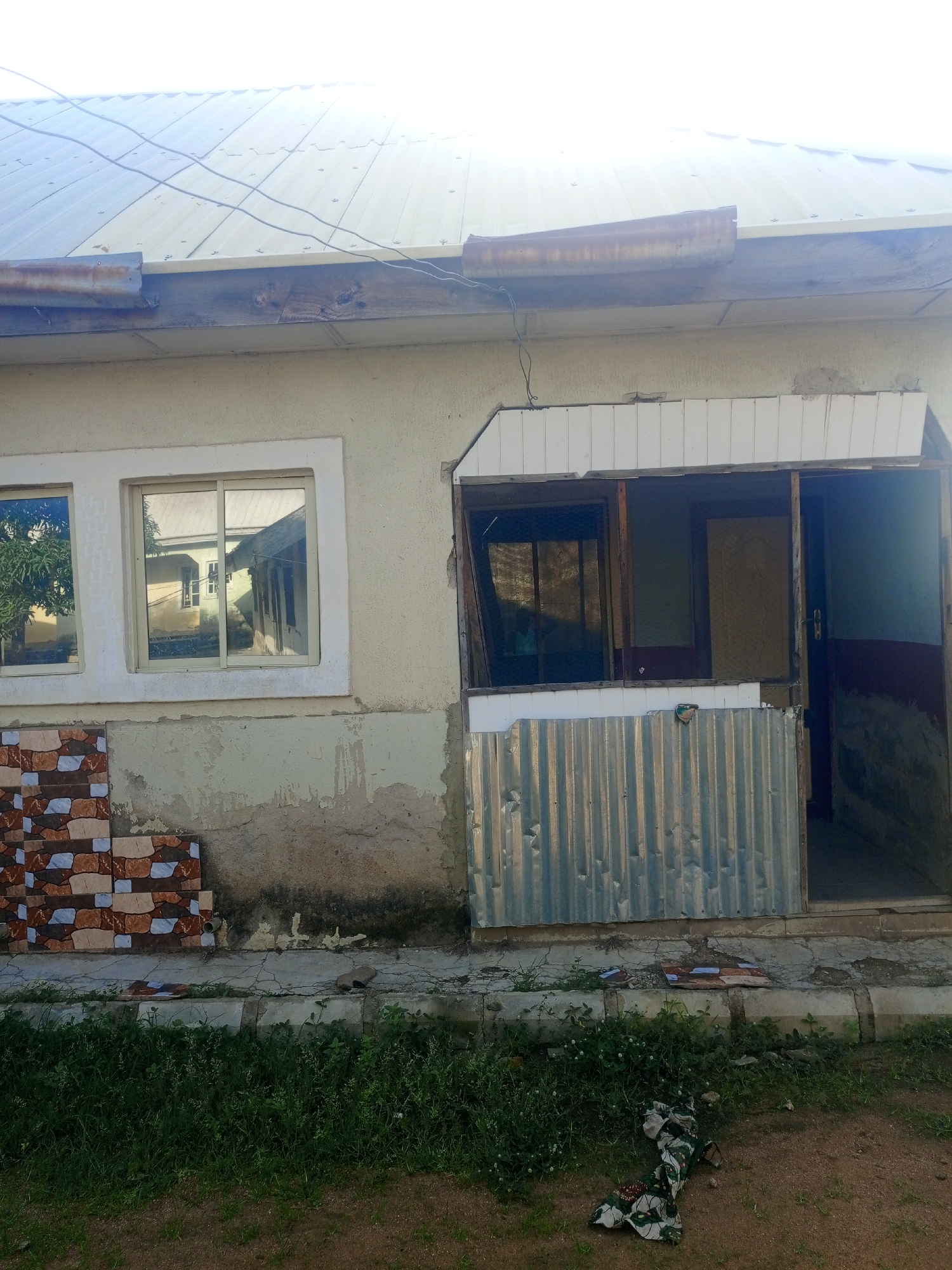 The image shows a dilapidated building in Jalingo, Nigeria. The building has a tin roof and a cream-colored facade. There are two windows on the left side of the building, and a covered entrance on the right. The entrance is partially obscured by corrugated metal sheeting. The building is surrounded by overgrown grass and dirt. There is some debris scattered around the entrance, including what appears to be a piece of fabric. The overall impression is one of neglect and disrepair.