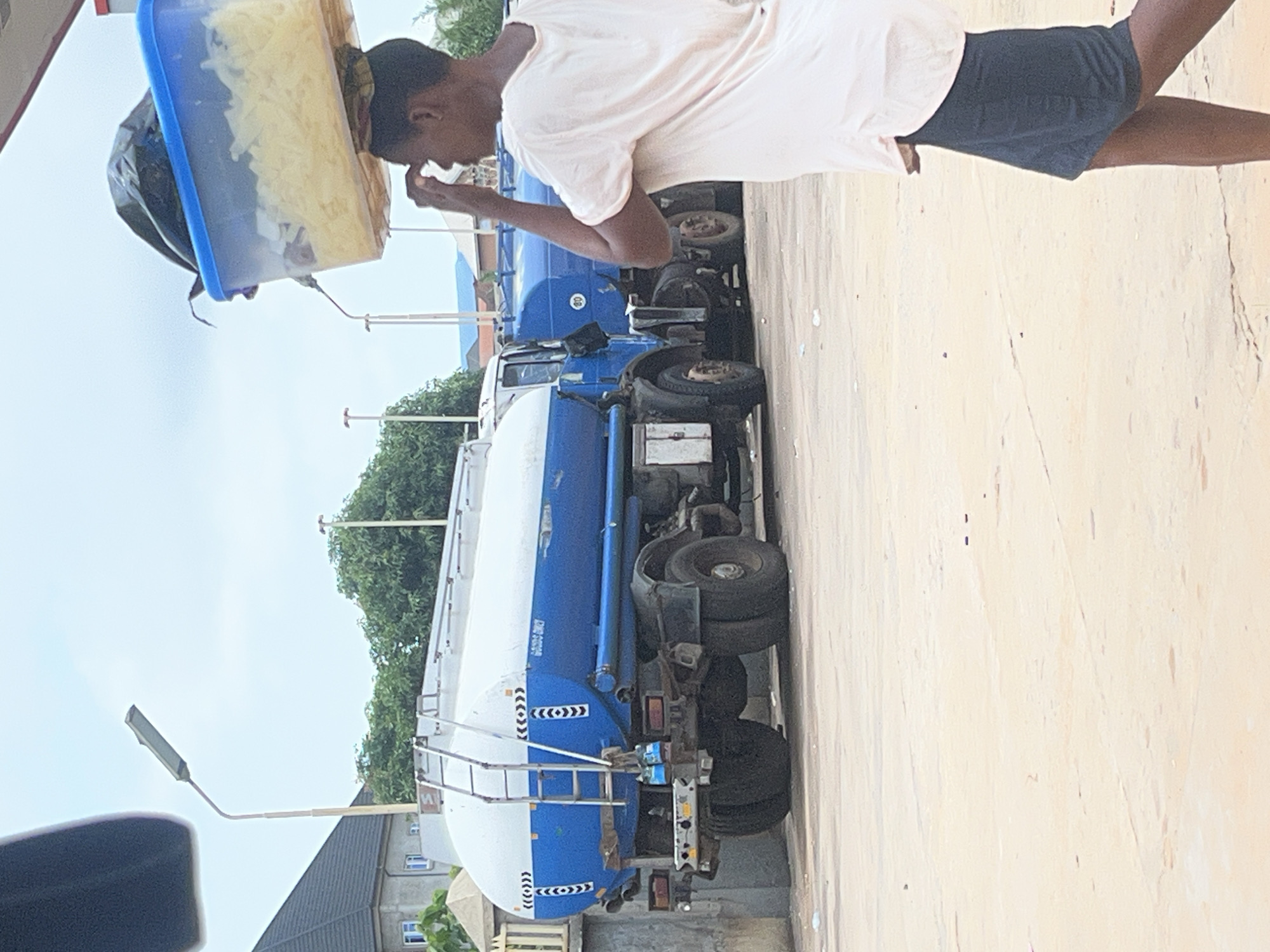 A man is seen carrying a clear plastic container on his head, with a black bag inside, standing on a paved surface. Behind him are two blue and white tanker trucks parked side by side. A street light and trees are visible in the background, along with a building with a dark roof. The location is an outdoor area in the city of Unknown, Unknown.