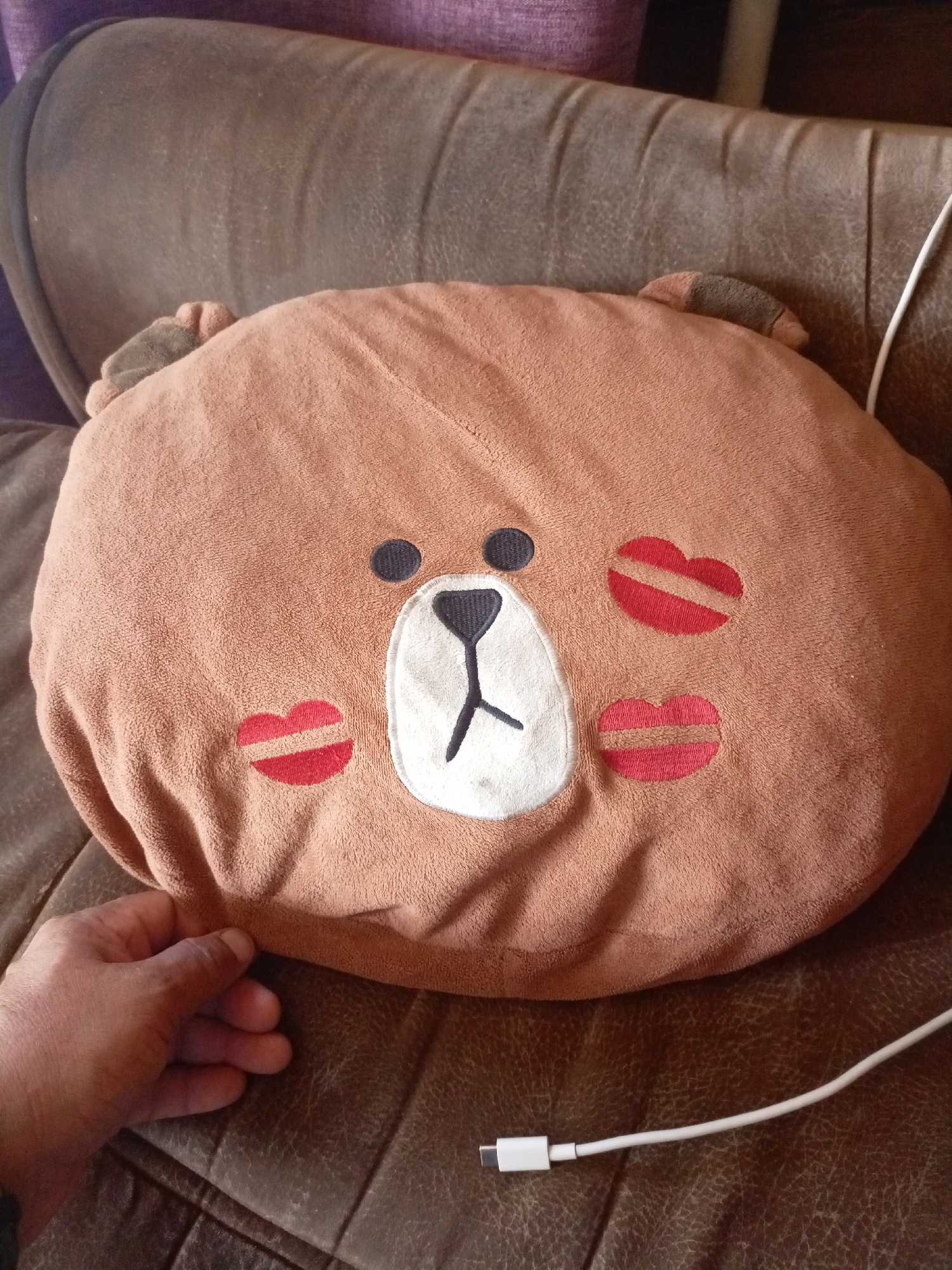 A brown, round plush pillow with a cartoon bear face design, featuring black eyes, a white muzzle with black stitching for a nose and mouth, and red lips with white stripes on either cheek, is placed on a brown leather sofa. The pillow has small brown ears on top. A human hand is visible on the lower left side of the image, near the edge of the pillow. A white cable with a white connector end is lying on the sofa to the right of the pillow. The background consists of the textured brown leather of the sofa and a purple upholstered headrest. There is no indication that this image is located in Churapcha, Russia.