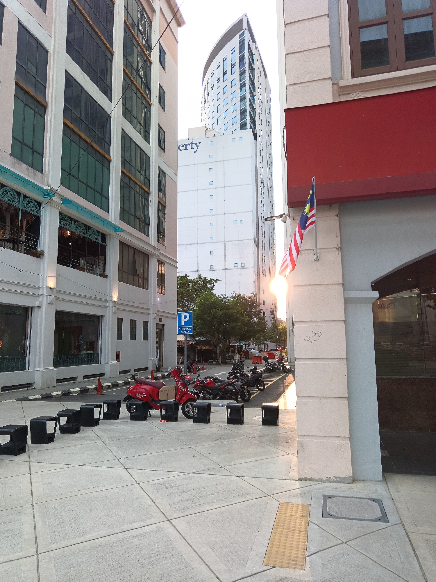 The image captures a street scene in Kuala Lumpur, Malaysia, characterized by modern architecture and urban elements. In the foreground, a paved walkway with distinct grid lines leads towards the viewer. Several black, modern-styled, cube-shaped benches are arranged in a line on the walkway. Beyond the benches, a bright red scooter is parked, facing left, with other motorbikes to its right. A blue "P" sign with "MOTORSIKA SAHAJA" written below it, indicating motorcycle parking, is visible behind the scooter.
The backdrop consists of various buildings. To the left, a multi-story building with large windows and white columns stands prominently. In the center, a tall, modern skyscraper with a curved top and many rectangular windows dominates the skyline. To the right, a section of a building with a red awning and a Malaysian flag attached to a white pillar is visible. The flag is partially illuminated by the setting sun, which casts a warm glow and creates lens flare. The sky is bright, suggesting either dawn or dusk. Trees are visible in the background between the buildings. The overall impression is a clean, urban environment with functional amenities like seating and parking.