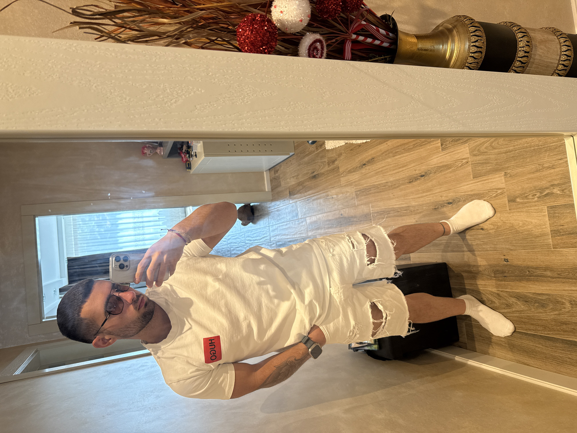 A man is seen in a mirror selfie. He is wearing a white t-shirt, distressed white shorts, and white socks. He has dark hair and a beard, and is wearing sunglasses. He is holding a smartphone. The reflection shows a room with a wood-grain tiled floor. In the background, there's a window with blinds and some furniture. Above the mirror is a decorative arrangement of branches, red and white ornaments, and a vase. There is no indication of the location being Gabrovo, Bulgaria.