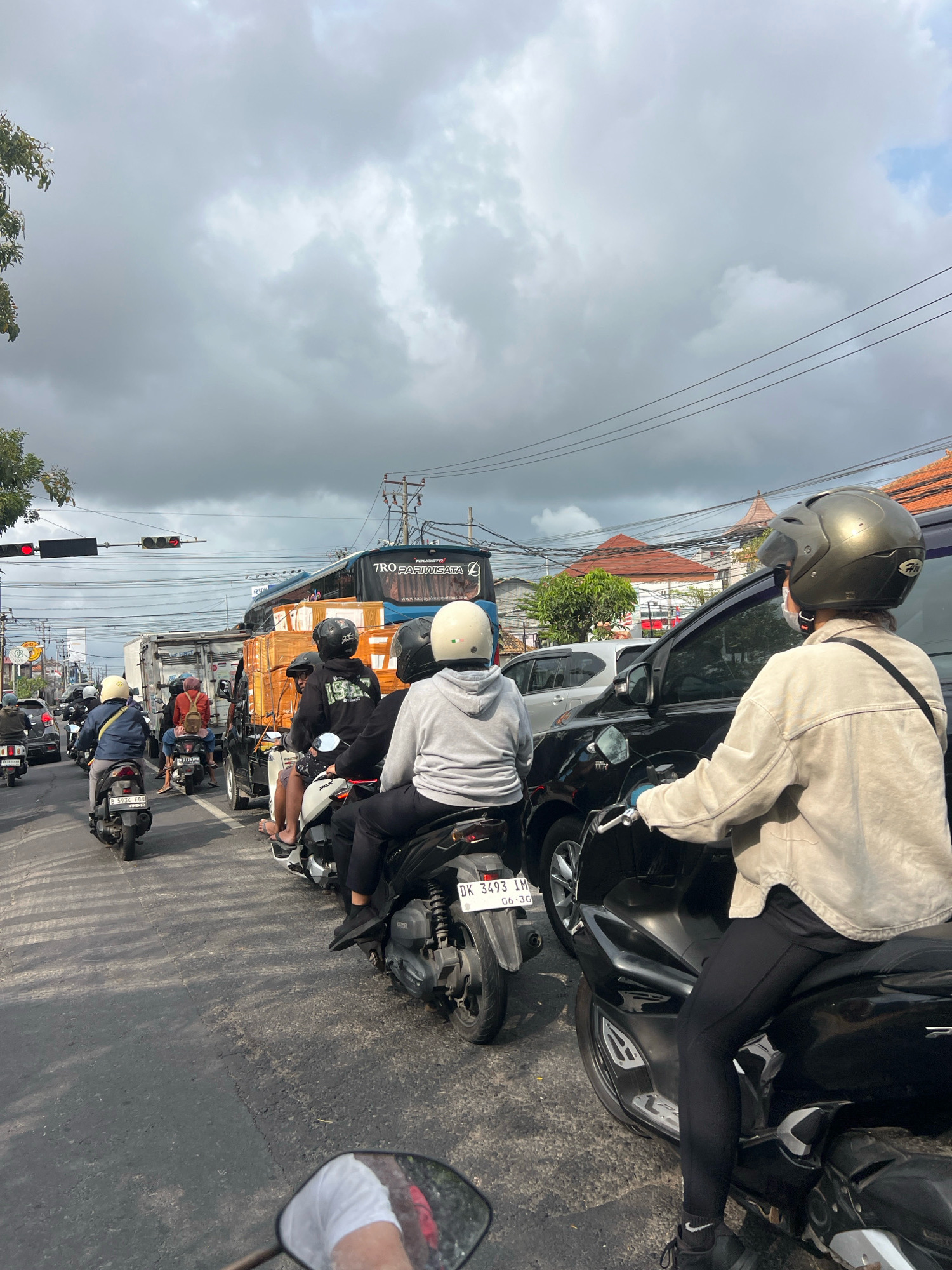 The image depicts a traffic jam on a city street in Denpasar, Indonesia, under a cloudy sky. Several motorcycles and a few cars are stopped on the road. On the right side of the image, a person wearing a beige jacket and a metallic helmet is riding a black motorcycle. The rider has a sling bag across their shoulder. In the middle of the frame, other motorcyclists are visible, some wearing helmets. Behind them, a white truck and a blue bus with the text "TRO PARIWISATA" are also stuck in traffic. The bus has the website "www.sanjayaekawisata.com" printed on it. Traffic lights are visible in the upper left corner, showing a red light. The road surface is asphalt with some cracks and shadows from overhead wires. Buildings with terracotta roofs are seen in the background, along with trees and utility poles.