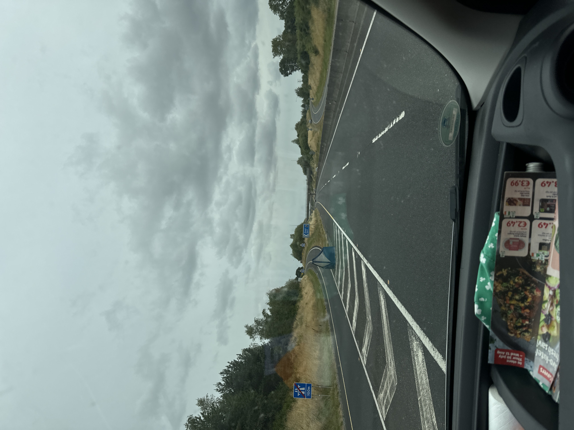 The image depicts a view from inside a vehicle traveling on a highway. The highway is paved and has multiple lanes, marked with white lines. On the left side of the road, there is a grass verge and a line of trees. A blue sign with a white symbol and "100m" is visible on the left. On the right side of the road, there is a concrete barrier and more trees. The sky is overcast with dark clouds. In the foreground, on the dashboard of the vehicle, there are some papers and a green cloth. The location is identified as Lackabeg, Ireland.