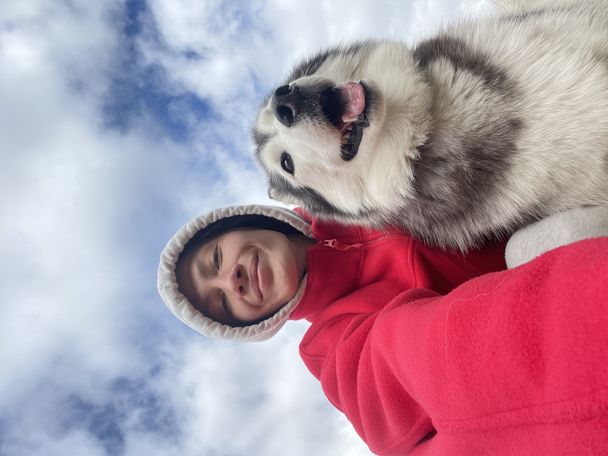 A low-angle, selfie-style photograph captures a person and a dog against a backdrop of a partly cloudy sky. The person, wearing a bright red fleece jacket with a white-lined hood, is positioned in the lower portion of the frame. The person is smiling slightly, with a nose ring visible. Above and to the right of the person is a husky dog, looking down with its tongue out. The sky is visible in the background, with patches of blue and white clouds. The location is not discernible as Izhevsk, Russia, or anywhere else specifically, based solely on the visual content of the image.