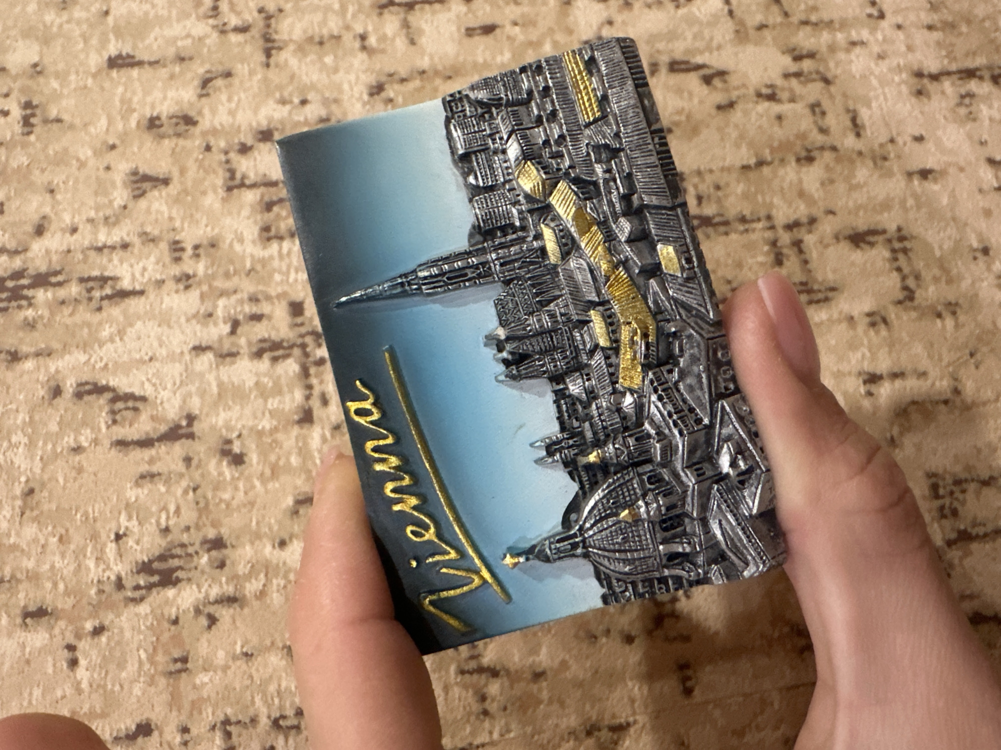 The image shows a hand holding a magnet. The magnet depicts a cityscape of Vienna, Austria, with the word "Vienna" written in gold lettering. The cityscape is rendered in detail, with silver and gold accents highlighting specific architectural features. The background of the magnet is a gradient of blue. The surface beneath the hand is a patterned beige and brown material.