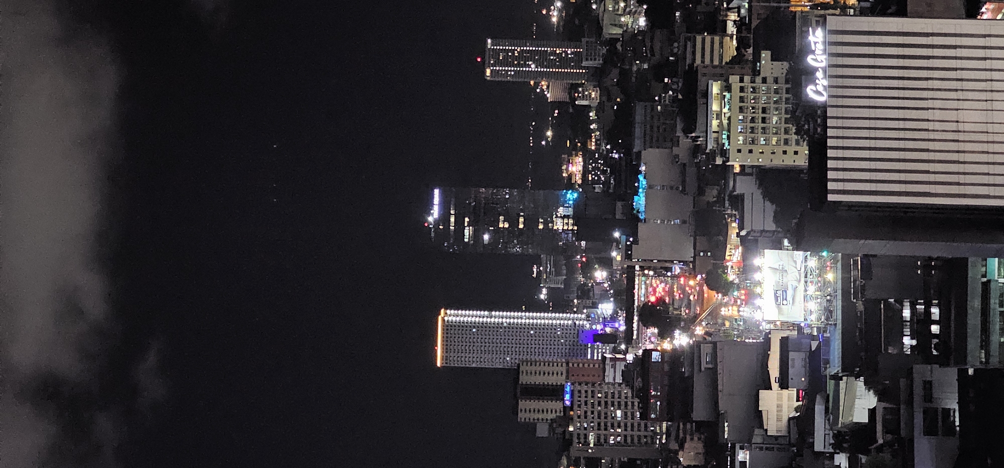 The image is a nighttime aerial view of the city of Makati, Philippines, captured on December 1, 2023, at 10:35 PM. The scene is dominated by a dark, cloudy sky with faint lights visible in the distance. In the foreground and midground, numerous buildings of varying heights and architectural styles are illuminated, creating a cityscape glow.
Prominent structures include a tall, slender skyscraper with a white facade and illuminated windows, topped with a bright orange band of light. To its right, another tall building features a facade of dark glass, with a grid of lit windows and a visible sign that appears to read "TRION SQUARE." Further to the right, a slightly shorter building also has a facade of illuminated windows.
The middle ground of the image shows a street level view of traffic, characterized by a multitude of red and white lights indicating moving vehicles. Buildings along this street are lit, with some displaying blue and white illuminated signage. A large billboard with a white background and text is also visible, illuminated from above.
The lower portion of the image reveals more buildings, including a building with a striped facade that has the illuminated text "Casa Grata" at its top. The overall impression is a bustling urban environment at night, with a dense concentration of illuminated structures. No people are discernible in the image.