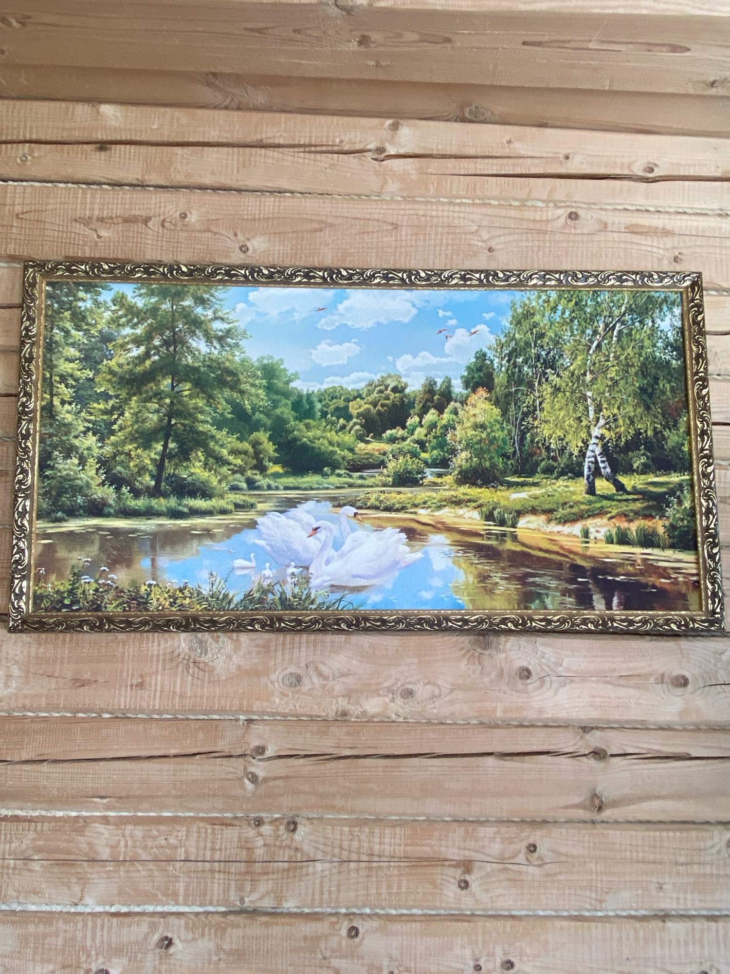 A framed painting of a serene lake scene with swans hangs on a wooden wall in Subash, Russia. The painting depicts a tranquil body of water surrounded by lush green trees and foliage. Two white swans are prominently featured in the foreground, gliding gracefully on the water's surface. The sky above is a clear blue with a few scattered clouds, and small birds are visible in flight. To the right of the scene, a lone figure, possibly a person, is depicted near a birch tree. The wooden wall itself is constructed from horizontal planks, showing natural wood grain and knots. The painting has a decorative, ornate gold frame.