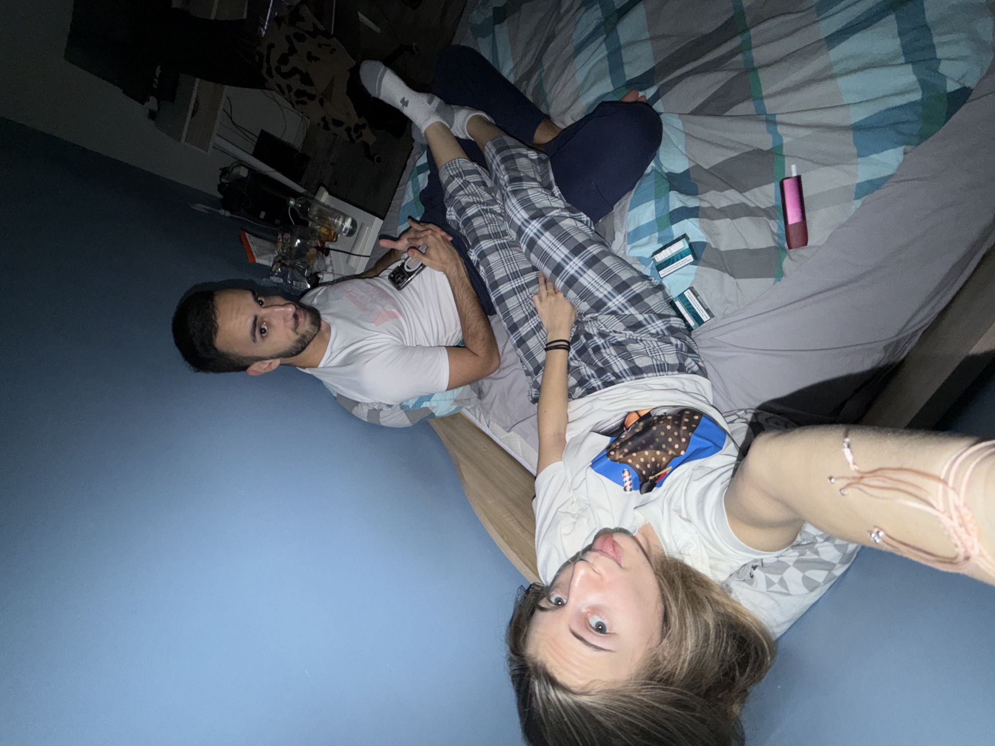 The image depicts two individuals lying on a bed with a patterned blue and white bedspread in a dimly lit room. A man with dark hair and a beard is wearing a white T-shirt and holding a phone. Next to him is a woman with blonde hair wearing a white T-shirt and plaid pajama pants. A pink vape and two packs of cigarettes are also on the bed. There is no evidence to determine the location as Plovdiv, Bulgaria.
