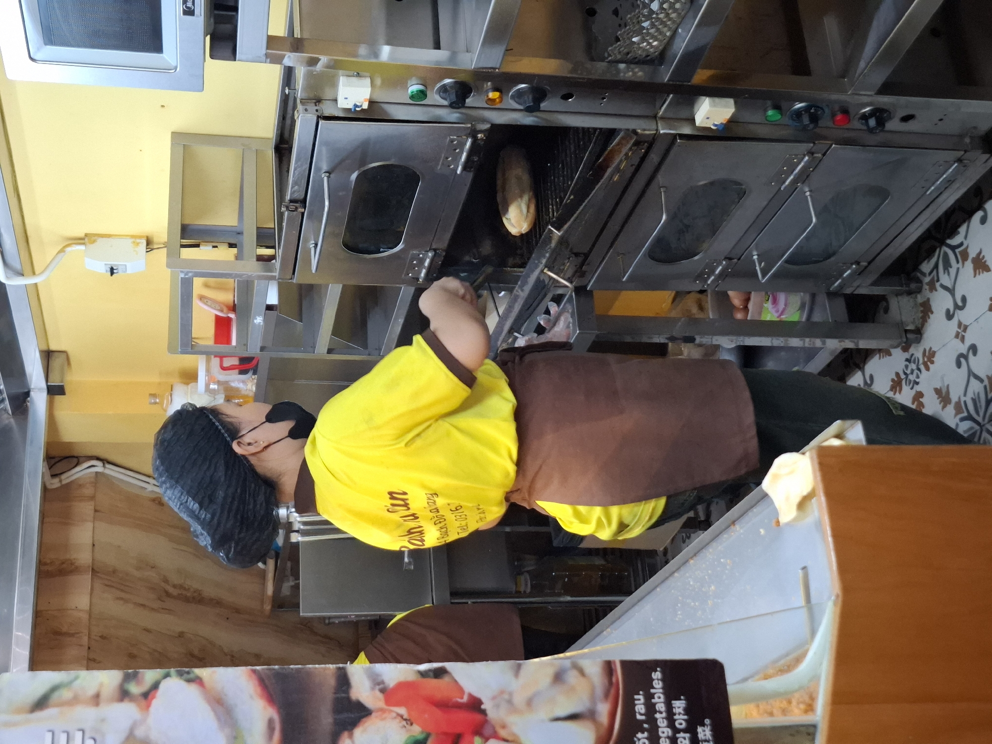 The image depicts an indoor scene within a food establishment, likely a bakery or sandwich shop, located in Nha Trang, Vietnam. A female employee wearing a yellow shirt, brown apron, and black hairnet is standing in front of a commercial double-deck oven. She is wearing a black face mask. The oven contains a loaf of bread on the upper rack, and the lower oven door is partially open. To the left of the employee, a menu board is visible, displaying text in English and Vietnamese. The environment includes stainless steel shelving, electrical outlets, and food preparation equipment. The floor is tiled with a decorative pattern. The overall impression is of a busy food service operation.