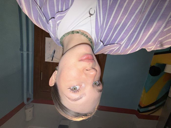 A rotated, close-up photo captures a light-skinned person with striking eyes. They are wearing a white t-shirt, topped with a lilac and white striped button-down shirt. A green beaded necklace adorns their neck, along with a silver crescent moon pendant necklace. Behind them, a partially visible door, a blue-painted wall, and a colorful object on the right are visible. The composition is slightly blurred due to the camera angle.