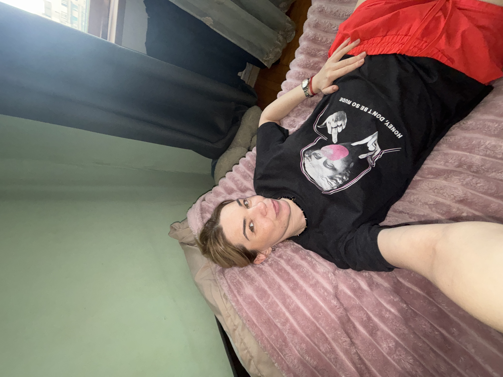 A young adult person is lying on a bed, angled on their back, and looking towards the camera. They have light brown hair, fair skin, and a neutral expression. They are wearing a black t-shirt with a graphic print and red shorts. A silver-toned watch is visible on their left wrist. The t-shirt print features a classical statue head blowing a pink bubblegum bubble, with two hands performing a gesture on either side. Upside down, the text "HONEY, DON'T BE SO RUDE" is visible on the shirt.
The bed is covered with a soft, pink, textured blanket and a light brown pillow. The room has light green walls. In the background, there's a window with bright daylight streaming through, illuminating a dark curtain that hangs beside it. A glimpse of buildings can be seen outside the window, suggesting an urban or suburban setting, consistent with the provided location context of Vsevolozhsk, Russia. The overall impression is casual and relaxed.