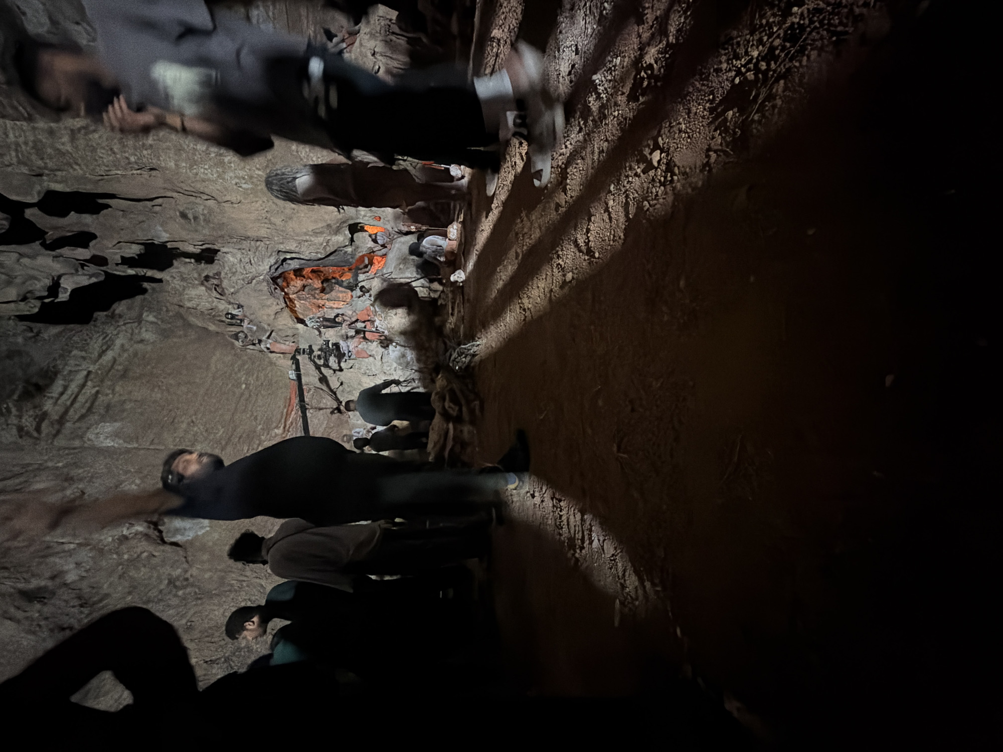 The image depicts a group of people inside a dimly lit cave, likely a tourist attraction, with the ground covered in sand and rocks. In the background, the cave walls are rough and textured, with stalactites hanging from the ceiling. Several individuals are visible, some standing and others sitting, appearing to observe or engage with the cave's features. The lighting is low, casting long shadows across the ground, with some areas illuminated by artificial light sources, possibly from flashlights or the ambient light of the cave's setup. There are also what appear to be decorative elements or religious figurines placed within the cave, some of which are illuminated with an orange glow. The overall atmosphere suggests a guided tour or a visit to a natural wonder. It is not possible to confirm the specific location as Guttapadu, India, based solely on the visual information provided in the image.