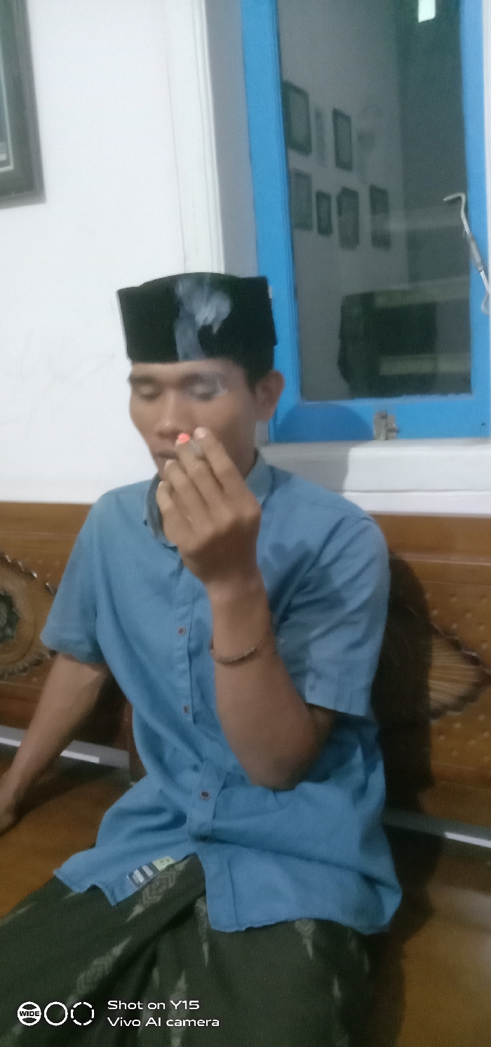 An adult male is seated indoors in what appears to be a residential setting in Brumbungan Kidul, Indonesia. He is wearing a black peci, a light blue short-sleeved collared shirt, and a dark green patterned sarong or trousers. A brown bracelet is visible on his right wrist. He holds a lit cigarette with a prominent red filter between the thumb and forefinger of his right hand, positioned near his mouth. Smoke is visibly emanating from the cigarette and dissipating around his head. His eyes are either closed or heavily lidded. The background features a white wall with a partially visible dark-framed object on the left. To the right, a bright blue window frame encloses a dark, reflective surface that shows indistinct reflections of multiple framed items. Below the subject, a brown wooden surface, possibly part of a bench or chair, is visible with some carved detailing on its left side.
