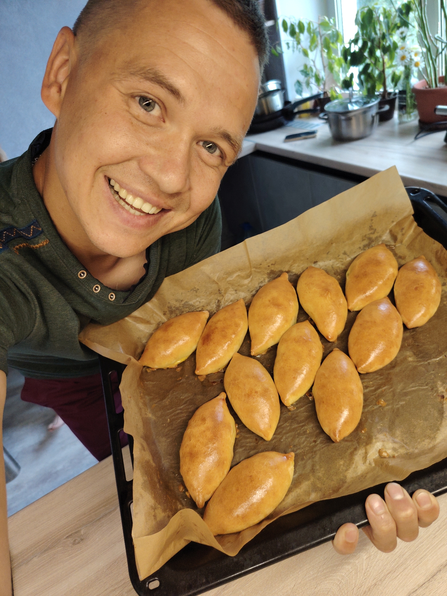 In a home kitchen setting, likely in Chaikovsky, Russia, a man with a shaved head and a green t-shirt smiles at the camera while holding a baking sheet filled with golden-brown, boat-shaped pastries. The pastries are arranged on parchment paper atop a black baking sheet. Behind him, a kitchen counter with various items including pots, a small pan, and plants in pots is visible. The man is the primary subject, and the pastries are prominently displayed. The overall scene depicts a domestic moment of culinary achievement.