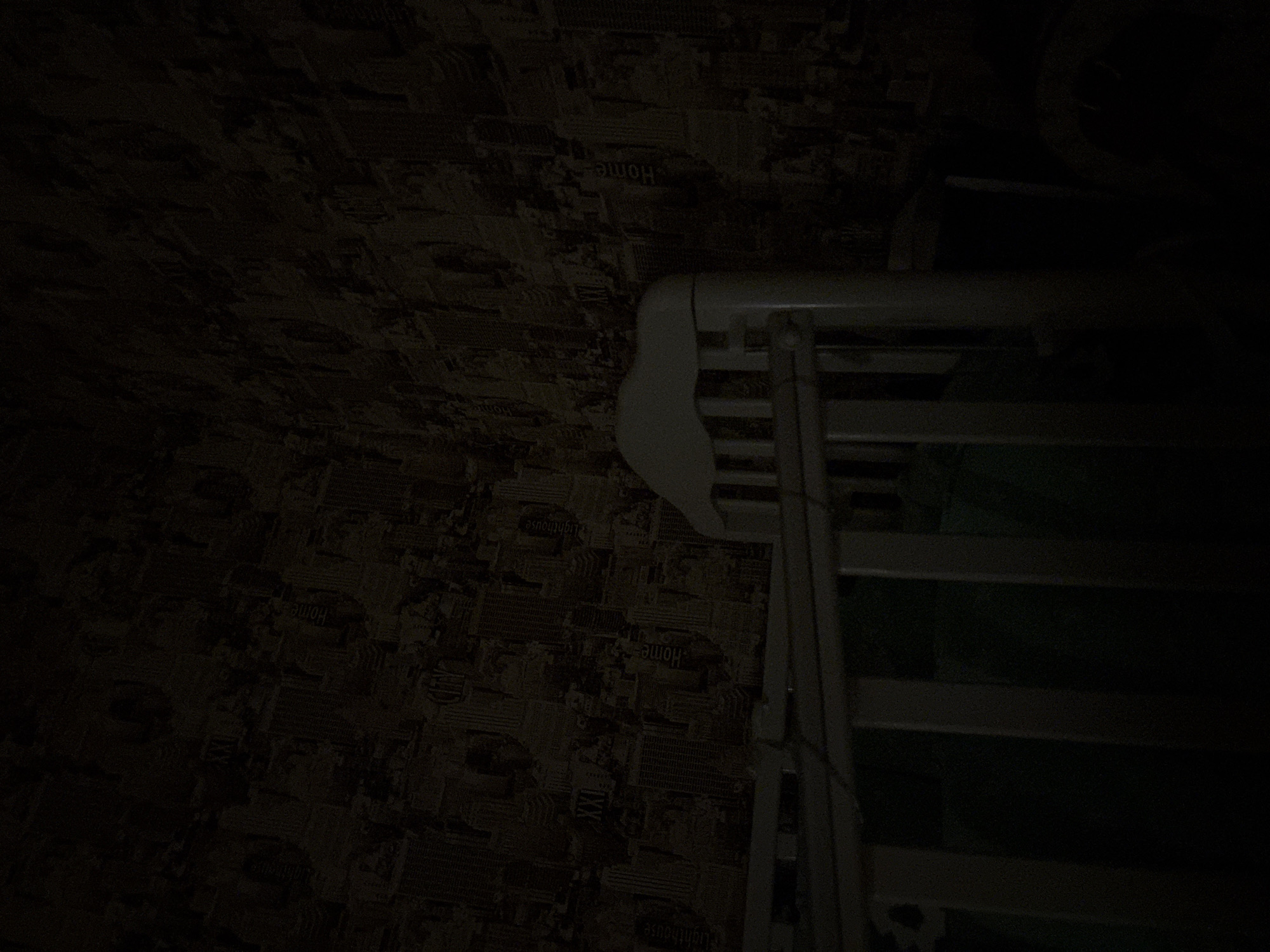 The image shows an indoor scene, possibly a bedroom or nursery, with a crib in the foreground. The crib is white with vertical slats and a curved headboard. The walls are covered in patterned wallpaper depicting a cityscape with skyscrapers and text that appears to be in English, including "CITY," "HOME," and "LIGHTHOUSE." The lighting is dim, casting long shadows and making it difficult to discern finer details of the room. There are no people or active events depicted in the image. The location is specified as Sovkhoza "Razdol'e", Russia, however, there is no visual evidence within the image to confirm this specific geographical location; the wallpaper design with English text suggests a possible non-Russian context or a deliberate thematic choice for the decor.