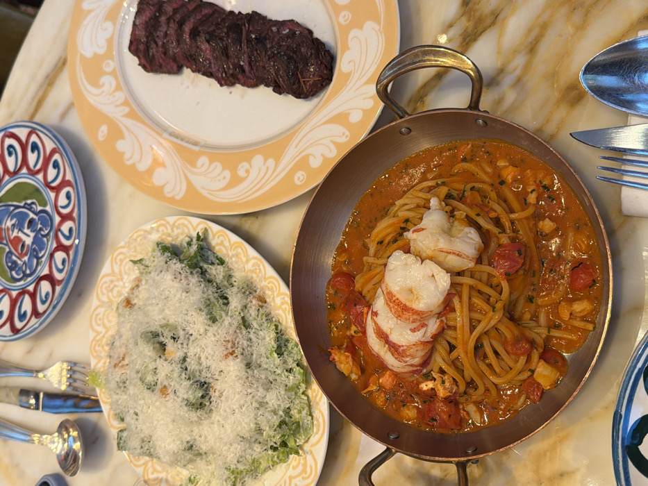 The scene depicts a high-angle view of a multi-course meal set on a polished, light-colored marble table, consistent with a high-end restaurant environment such as Cipriani in the Dubai International Financial Centre (DIFC), Dubai, United Arab Emirates.
In the foreground and center, a round copper pan contains spaghetti served in an orange-red sauce with diced tomatoes and herbs, topped with two large, cooked lobster tails. To the immediate left of the copper pan, an oval white plate with a yellow scroll-patterned rim holds a Caesar salad heavily garnished with grated white cheese. To the upper right, another oval white plate, this one with an orange scroll-patterned rim, displays several slices of medium-rare steak, seasoned with visible coarse salt. Various pieces of polished silverware, including forks, knives, spoons, and a small ladle, are scattered on the marble surface. A small, round decorative plate with blue, red, and white patterns is positioned in the upper center. No individuals are present in the image.