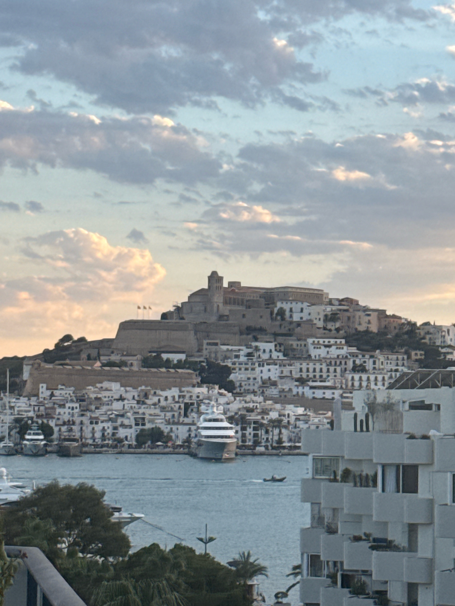 A wide shot captures the city of Ibiza, Spain, with a view of the Dalt Vila, a fortified old town, perched atop a hill. The Dalt Vila's ancient stone walls and buildings are visible, topped by a prominent structure that appears to be a cathedral or castle. Below the old town, a dense collection of white-washed buildings with red-tiled roofs cascades down the hillside towards the sea.
In the foreground, a modern, white residential building with a unique, stepped facade and balconies is partially visible on the right side of the frame. The sea in front of the city is dotted with several boats, including a large white yacht anchored in the harbor, and a smaller motorboat moving across the water. The sky above is filled with a mix of white and grey clouds, with patches of blue sky peeking through, suggesting either sunrise or sunset. The overall impression is of a coastal city with a rich historical past coexisting with modern development.