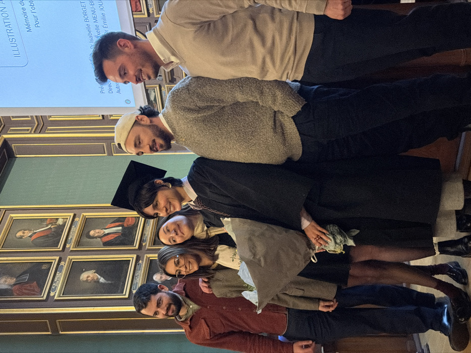 A group of six individuals are lined up, seemingly posing for a photo indoors. The first two men are wearing casual attire like a white shirt, grey sweater, and a cap. A woman in a graduation gown and cap follows. Two more women stand below her, one wearing glasses. The last person, a man, is in a red jacket. On the left, a wall is adorned with several framed portraits. A projection screen in the upper left displays text, including "ILLUSTRATION A," "Isabelle BORGET," and "Émilie JOU."