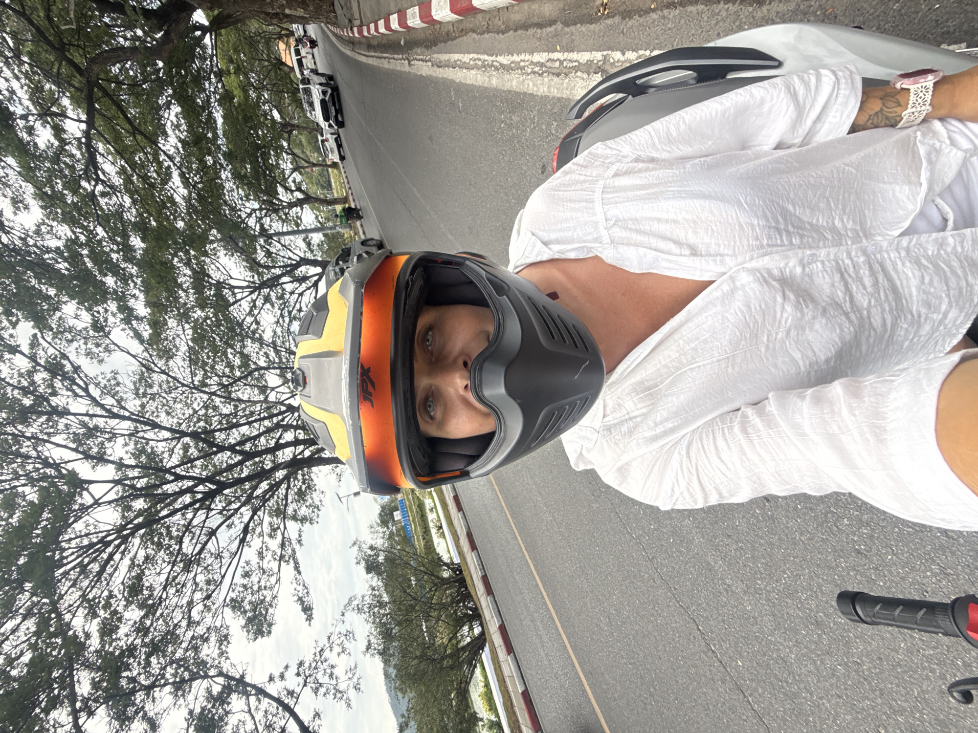 A woman wearing a white shirt and a full-face motorcycle helmet with yellow and orange accents is riding a motorcycle on a paved road. The road has a curb with red and white markings on the right side. Trees with dense foliage line the right side of the road, and a body of water is visible on the left. Other vehicles, including cars and motorcycles, are present on the road. The sky is overcast. The image appears to be taken in a tropical or subtropical environment, likely in a city or town within Ko Samui District, Thailand.
