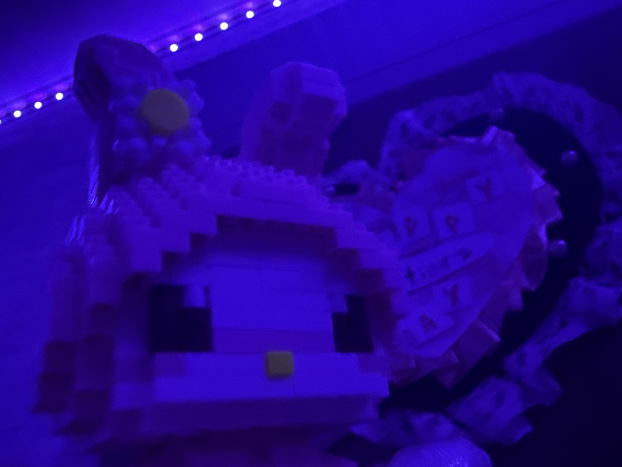 A pink, pixelated figure resembling a cartoon character is illuminated by purple lighting. The figure appears to be constructed from small, interlocking blocks. It has a large, square head with dark rectangular eyes and a small, square yellow nose. The figure has two tall, pointed ears and a circular yellow embellishment on one side of its head, possibly a flower. To the right of the figure, a heart-shaped object made of white lace and fabric is visible, with pieces of paper attached that have letters on them, partially obscured by the purple light. The background is dark, with a string of small, white lights running along the top of the frame, suggesting an indoor setting, likely a room in Rayevskiy, Russia. The lighting creates a low-visibility, high-contrast environment.