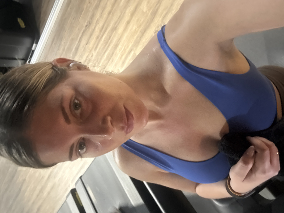 An adult female is depicted indoors, likely within a gym environment. She has light-toned skin, light brown hair pulled back, and appears visibly sweaty on her face, neck, and shoulders. She is wearing a royal blue sports bra, with a segment of dark brown attire visible beneath it. A white earbud is present in her right ear, and a dark-colored wristband is on her left wrist. She holds a dark, crumpled towel with both hands near her chest, below the sternum. Her gaze is directed straight forward, consistent with taking a selfie. In the background to her left, two treadmills are visible, featuring light-colored running decks and dark grey frames. The flooring consists of light-colored wooden planks. The upper right corner reveals a dark, rectangular piece of gym equipment and a reflective surface, possibly a mirror. The specific location within the city of Paris, France, cannot be determined from the visual evidence provided in the image.