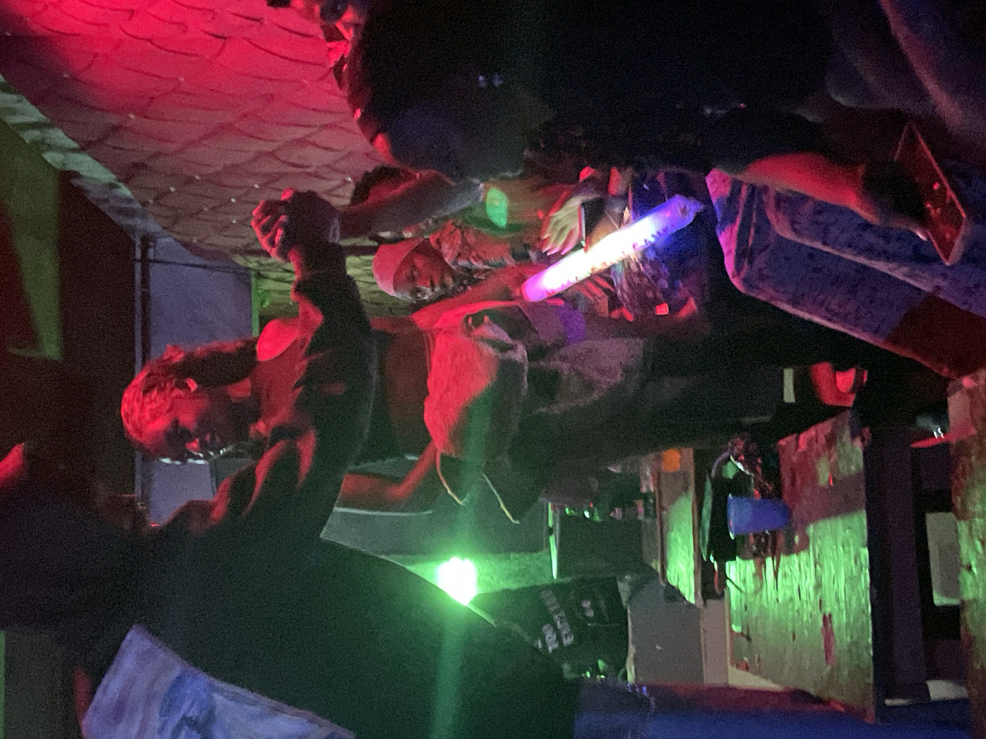 In Durumi, Nigeria, a dimly lit indoor space, possibly a club or bar, features several people interacting. In the foreground, a person in a dark top and light-colored bottoms is seen from the side. To their right, a woman with long, dark, curly hair, wearing a dark top and a red skirt or fabric around her waist, is dancing with someone. Her hands are raised. In the background, to the right, a woman in a headscarf and patterned clothing looks towards the camera, holding a glowing stick. Another person's hand is visible holding a cell phone. Tables with bottles and a blue cup are visible in the foreground. The environment is illuminated by vibrant red and green lights, creating a festive and energetic atmosphere.