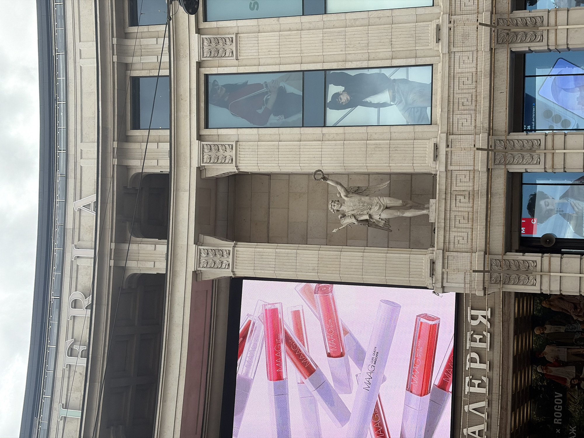 This image captures a vertically oriented view of a multi-story building facade, characteristic of classical architecture, likely in Saint Petersburg, Russia, based on visible text. The building features light-colored stone or stucco, ornate decorative elements, columns, and large windows.
A prominent architectural detail is a sculpture of an angel-like figure, possibly Icarus or another mythical character, holding a laurel wreath, situated within a recessed niche.
The facade is extensively utilized for large digital advertisements. A large screen in the lower section displays various cosmetic products from the brand "MAAG," including lip glosses and a mascara tube labeled "CURL QUEEN MASCARA," with shades of pink, red, and white packaging.
Further up, several window-like screens showcase different content: one displays two models, one in a red outfit and another in a black top; another shows a close-up of a person's face; and a third exhibits a smartphone screen with blue/purple graphics and the partial Cyrillic text "нный интеллект" (part of "artificial intelligence") along with "26." A narrower screen on the right side features a group of people, possibly models in historical or elaborate attire, accompanied by the text "X ROGOV" and a Cyrillic "Ж".
Large Cyrillic letters spelling "ГАЛЕРЕЯ" (GALEREYA), meaning "Gallery," are affixed to the building's upper section, indicating it might be a shopping gallery or a major retail establishment.
The sky is overcast, suggesting daytime with diffused light. The scene depicts a modern commercial setting integrated into a historically styled building, highlighting advertising and consumer goods.