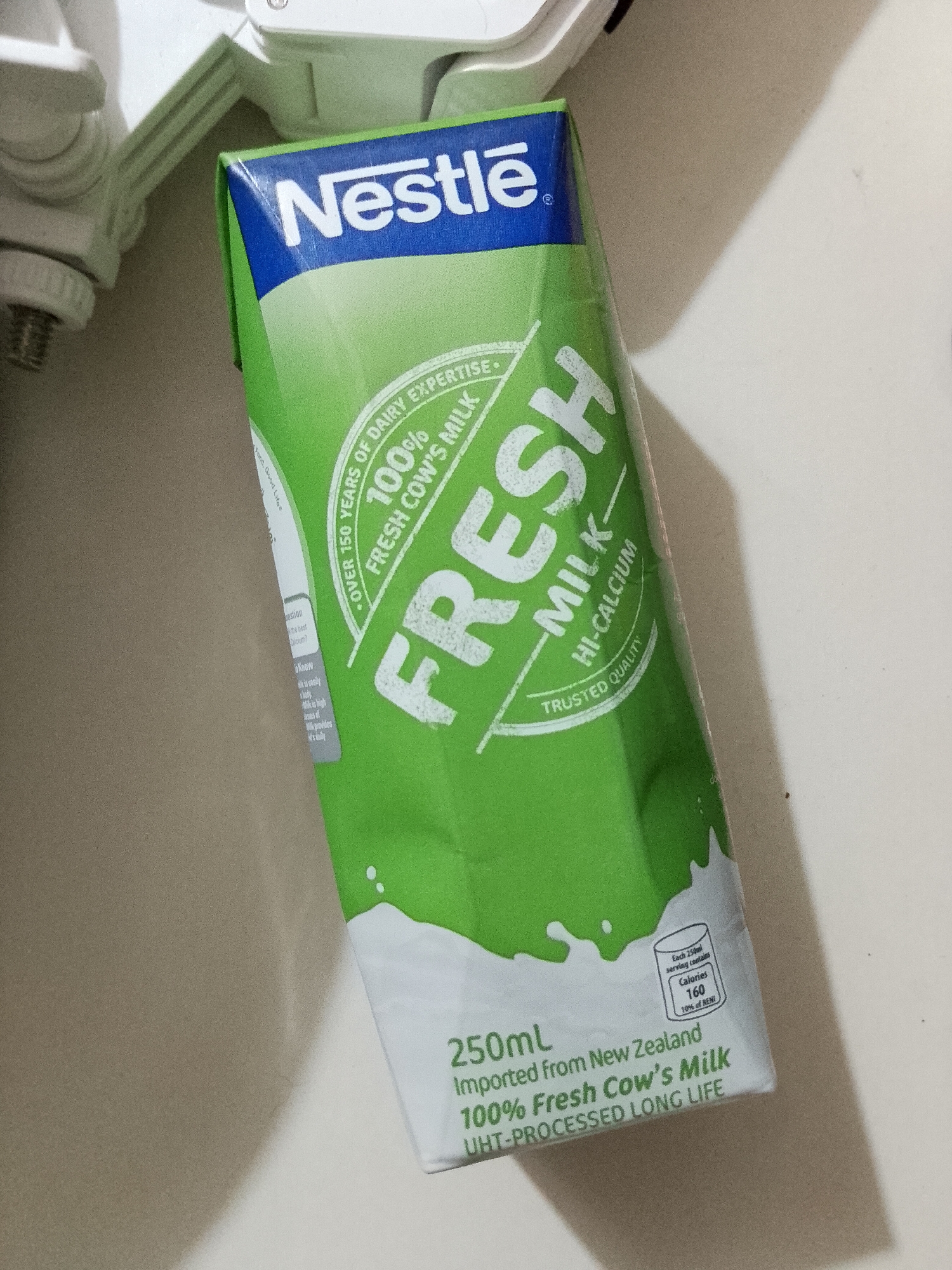 A carton of Nestle Fresh Milk, primarily green and white, is positioned diagonally on a light-colored surface. The carton's top features a blue band with the "Nestlé" logo in white. The main body of the carton is bright green, bearing the large white text "FRESH MILK" and "HI-CALCIUM."
Key information visible on the carton includes:
- "OVER 150 YEARS OF DAIRY EXPERTISE"
- "100% FRESH COW'S MILK"
- "TRUSTED QUALITY"
- Volume: "250mL"
- Origin: "Imported from New Zealand"
- Processing: "UHT-PROCESSED LONG LIFE"
- Nutritional information per 250ml serving: "Calories 160" and "10% of RENT" (likely Reference Nutrient Intake).
- Other partial texts include "Food. Good Life" and fragments of a Q&A section like "question," "best," "know," and "Calcium?".
The carton's design incorporates a white splash graphic at the bottom. The background consists of a light, possibly off-white, surface, which appears to be a tabletop or counter. In the upper left corner, parts of white plastic or metallic objects are visible, casting shadows on the surface, indicating indoor lighting. There are no people or specific events depicted. The image was captured in Davao City, Philippines.