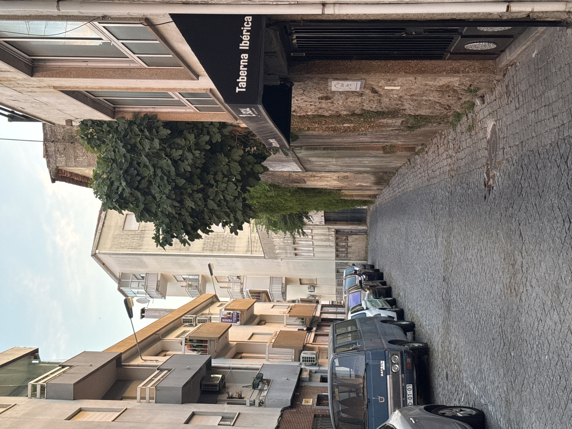 A narrow cobblestone street in Castelo Branco, Portugal, is lined with parked cars and buildings. On the left, residential buildings with balconies and air conditioning units are visible. The street features a blue Datsun van in the foreground, followed by a line of other cars. On the right, a stone wall with ivy and a building with a black awning reading "Taberna Ibérica" are prominent. The sky above is a pale blue with some clouds, suggesting it is either early morning or late afternoon. The overall impression is of a quiet, urban street scene.